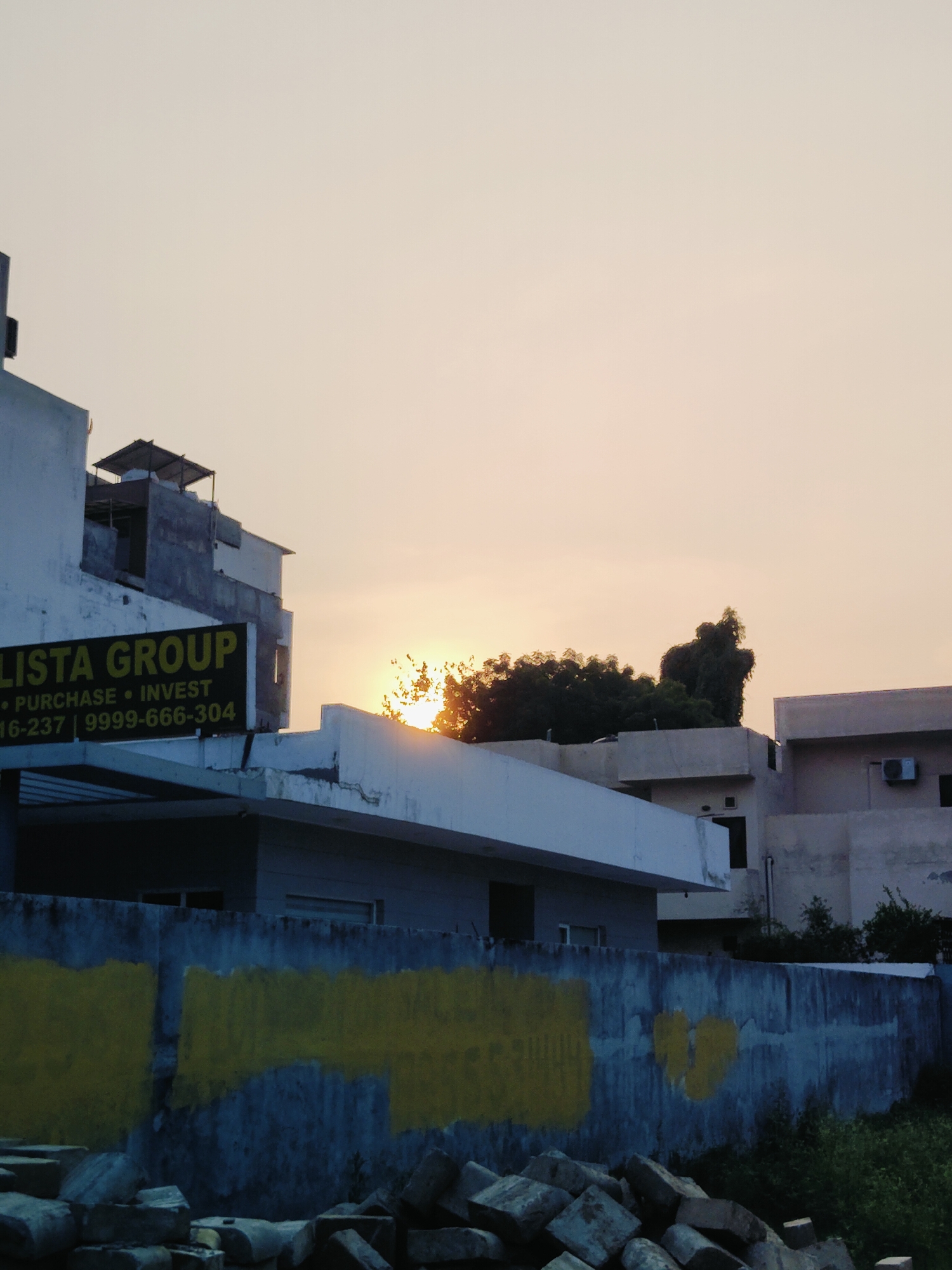 The image shows an outdoor scene at sunset in Gurgaon, India. In the foreground, there is a concrete wall with a blue and grey textured finish, marked with two large, roughly painted yellow rectangular shapes. Piles of broken concrete blocks and rubble are stacked in front of the wall. Behind the wall, a multi-story building is visible, with a lower section painted white and a darker upper section. A sign on the white section reads "LISTA GROUP" in yellow text, followed by "PURCHASE • INVEST" and contact numbers. Further in the background, more buildings with light-colored facades are visible. Trees with dark green foliage are present, and the setting sun casts a warm glow through the branches. The sky is a pale orange-yellow, indicating the time of day. There are no people visible in the image. The overall impression is of an urban or semi-urban environment during twilight.