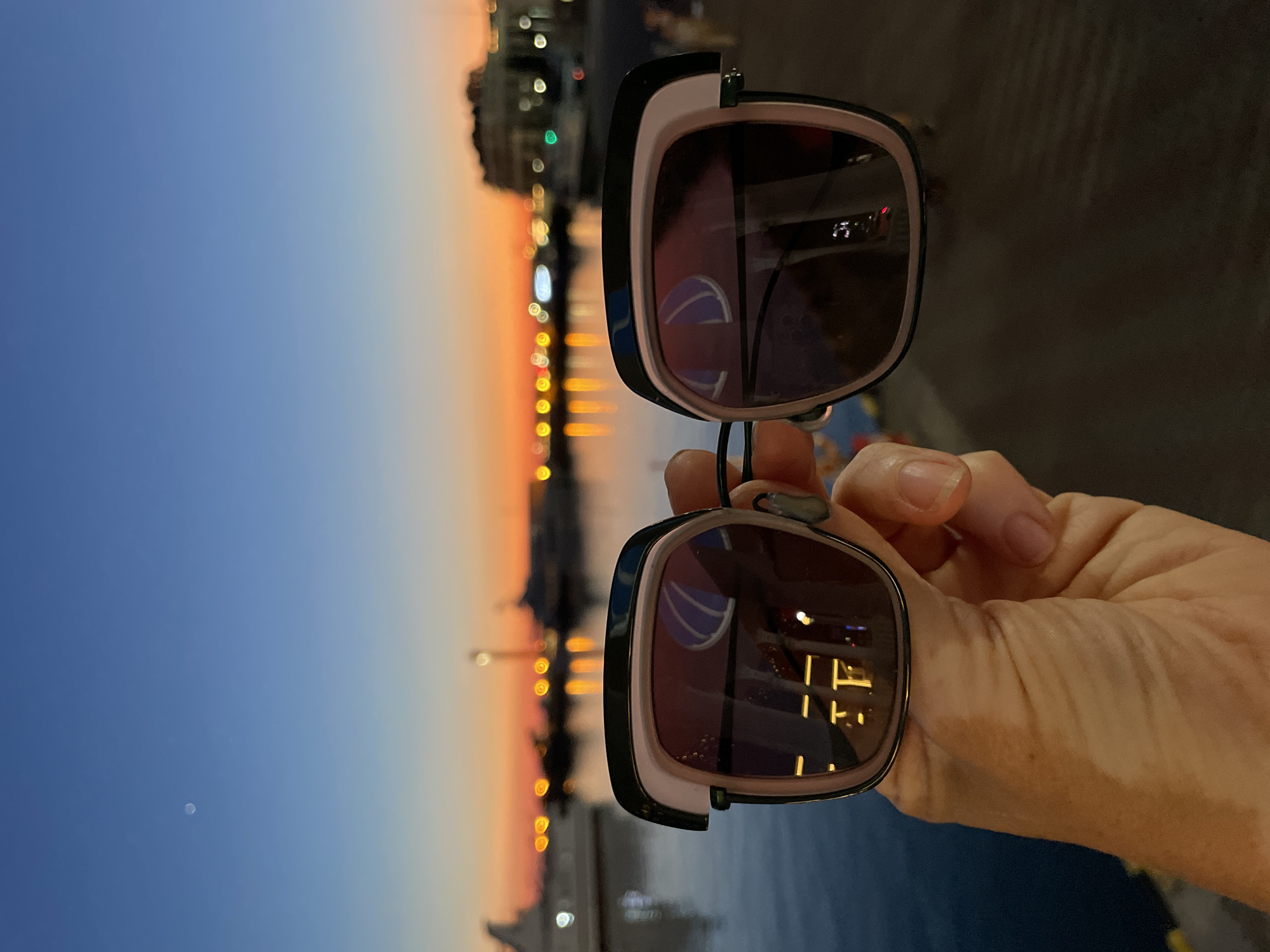 A pair of sunglasses is held up in the foreground, obscuring a person's face. The sunglasses have thick black and pink frames and dark lenses. The lenses reflect a dimly lit scene with glowing lights, possibly from buildings or streetlights, and a hint of a person's face. In the background, a body of water is visible, and beyond that, a skyline with illuminated buildings and structures is silhouetted against a twilight sky. The sky transitions from a deep blue at the top to shades of orange and yellow near the horizon, indicating that it is either dawn or dusk. The overall mood is serene, with a sense of an evening or early morning by the water. The location is identified as Sochi, Russia, suggesting a coastal setting.