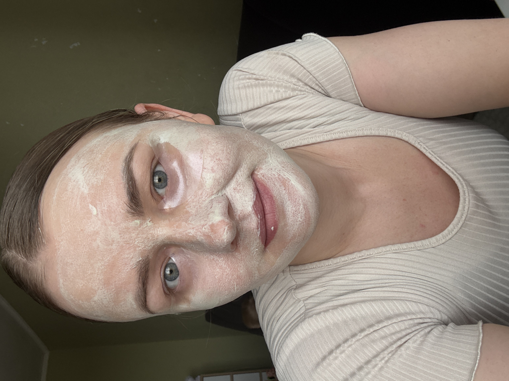 A woman is depicted in a close-up shot, with her face oriented towards the viewer. A light-colored cream or mask is applied to her face. She has fair skin, and her eyes are light colored. She's wearing a beige, ribbed short-sleeved shirt. The background is out of focus. Based on the prompt's context, the location is Chaikovsky, Russia, but this cannot be confirmed from the image alone.