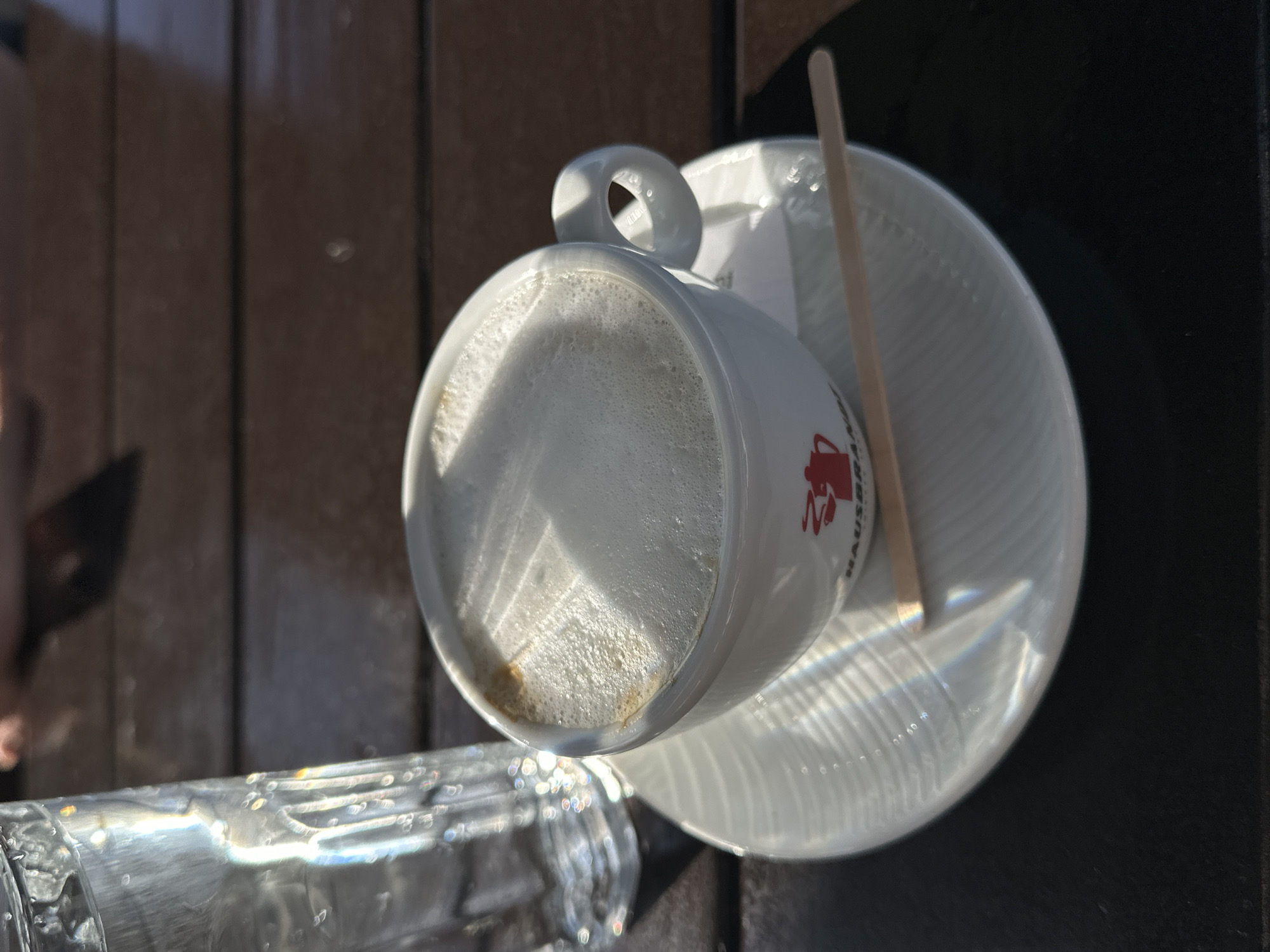 A close-up, high-angle shot captures a partially consumed cappuccino in a white "Hausbrandt" branded cup, positioned on a white saucer with a wooden stirrer. The cup and saucer sit on a dark brown surface next to a partially visible glass of water. The cappuccino features a frothy top with light and dark brown patches. The setting appears to be outdoors, likely a cafe or restaurant in Šušanj, Montenegro, suggested by the European brand name. The scene is bathed in bright sunlight, evident from the prominent reflections on the saucer and glass. A portion of a light-colored wooden surface is also visible in the background.