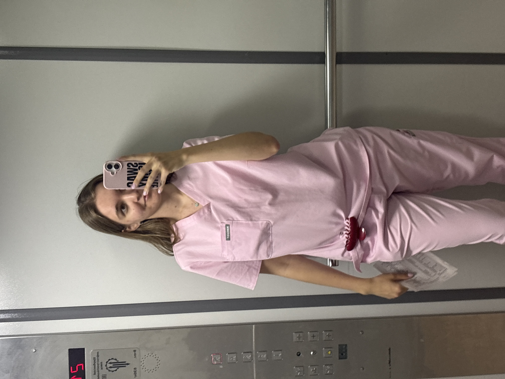 A young woman is captured in a mirror selfie, her image rotated 90 degrees counter-clockwise. She is wearing a light pink medical scrub uniform, consisting of a v-neck top with a chest pocket and matching pants. A red hair claw clip is attached to her waistline. Her left hand holds a folded piece of paper, while her right hand holds a light pink smartphone, used to take the photo. Her phone case displays mirrored black text that reads "SWANKY VIBES". Her hair is light brown and falls to her shoulders, and she wears a subtle necklace and an earring.
The setting appears to be inside an elevator, reflected in its mirror-like walls. The walls are a light gray, with a horizontal dark gray strip running across the middle and a vertical metallic bar on the right side. The elevator control panel is visible at the bottom of the image. A digital display on the panel shows "05", indicating the fifth floor. Below this, there is an "OTIS" logo and Cyrillic text, likely "Отдел технического обслуживания" (Technical Service Department) and "Диспетчерская" (Control Room/Dispatch). The panel also features numerous square buttons for different floors, with some appearing illuminated, as well as door control and alarm buttons. A small tag with the mirrored text "TOSSIN" is visible on the left chest pocket of her scrub top. The presence of Cyrillic text on the elevator panel aligns with the provided location context of Perm, Russia.