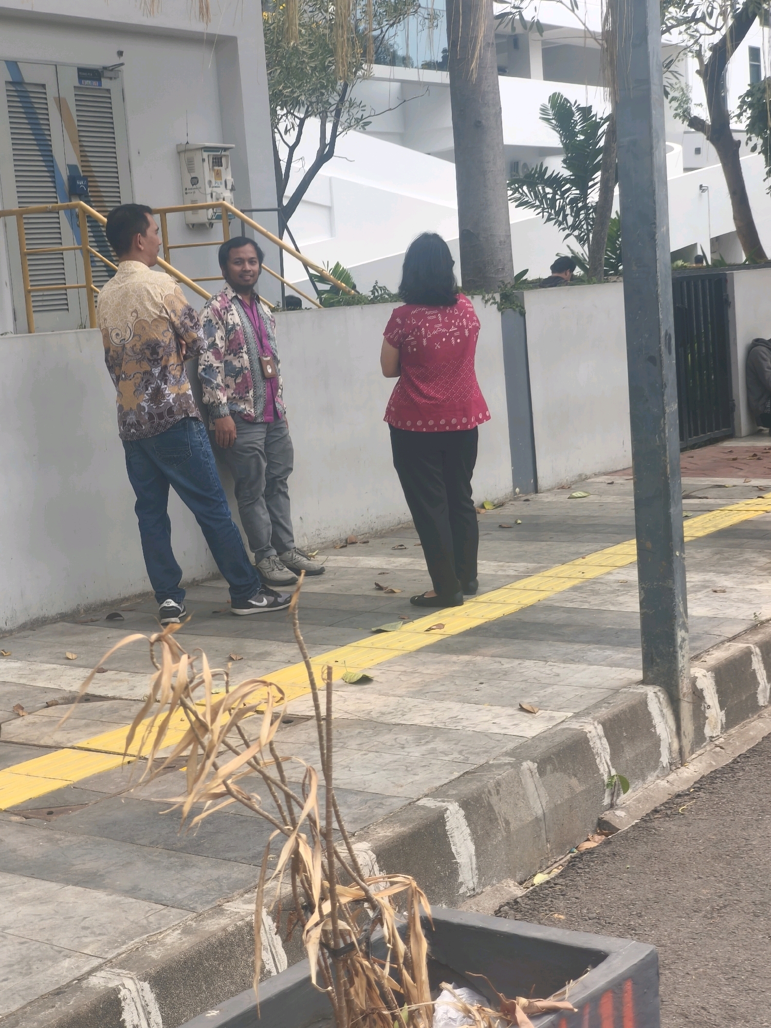Three individuals are standing on a sidewalk next to a white building with a yellow handrail and a blue stripe on a door. Two men, on the left, are facing each other and are both wearing patterned shirts. The man on the far left wears a tan and brown patterned shirt and blue jeans. The man to his right wears a floral patterned shirt and grey pants. A woman, on the right, is facing away from the camera, wearing a pink patterned shirt and black pants. A yellow tactile paving line runs along the edge of the sidewalk. Dead plant material is visible in the foreground. A dark grey pole stands to the right of the woman. The setting appears to be a public area in East Jakarta, Indonesia, indicated by the architecture and street elements.
