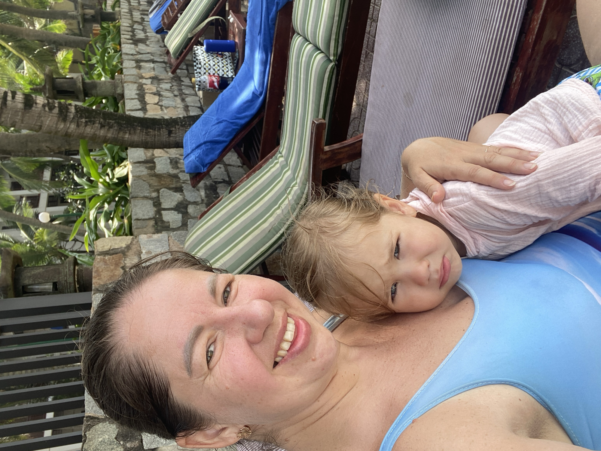 A woman and a young child are seated on lounge chairs at a resort in Phu Thuy Ward, Vietnam. The woman, wearing a blue swimsuit, smiles at the camera while holding the child, who is looking away with a pensive expression. Behind them, lounge chairs are arranged near a stone wall adorned with plants and palm trees, suggesting an outdoor tropical setting. The overall scene depicts a relaxed moment at a holiday destination.