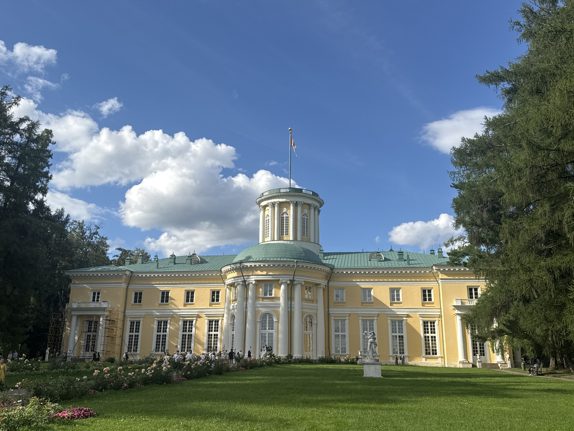 The image shows the main palace of the Arkhangel'skoe estate, located in Russia. The palace is a large, yellow, neoclassical building with a green roof. It has a central rotunda topped by a dome and a flag. The palace is surrounded by a well-maintained lawn, flower beds, and tall trees. Several people are visible on the grounds, some walking and others sitting. One person is on a bench to the right, and a group is gathered in front of the palace on the left. A statue of two figures stands on a pedestal in the foreground, and scaffolding is present on the left side of the palace, indicating possible ongoing restoration work. The sky is blue with scattered white clouds, suggesting a pleasant day.