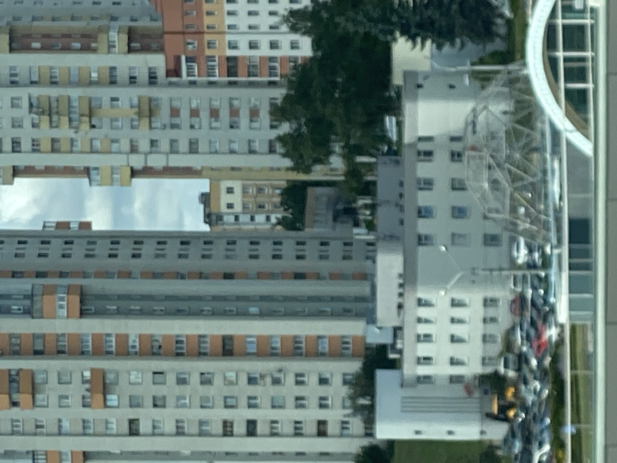 The image shows a view of high-rise residential buildings in Minsk, Belarus. In the foreground, there are lower-rise buildings with a geodesic dome structure. A parking lot filled with cars is visible in front of these buildings. The sky is overcast. The image is taken from a distance, likely from another building or elevated vantage point.