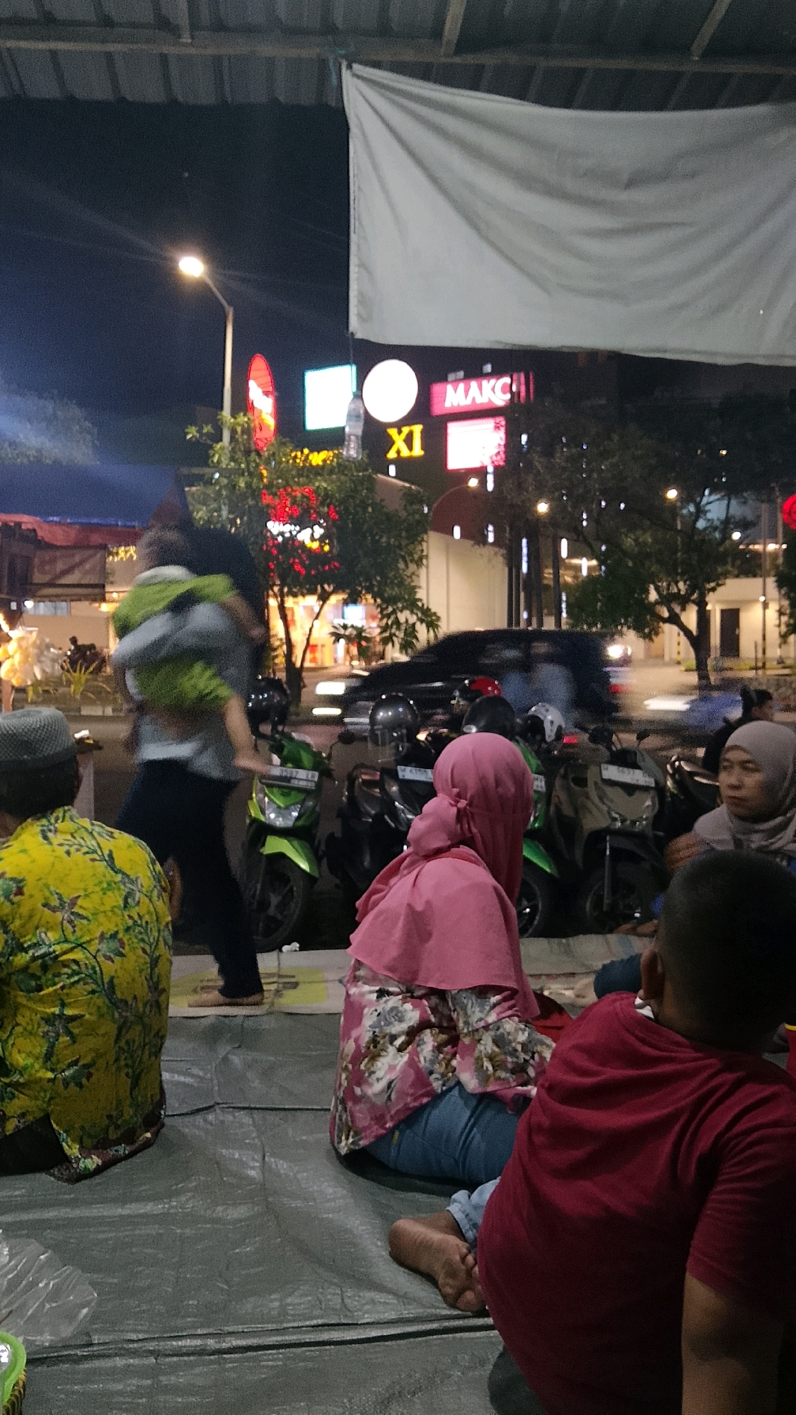 The image depicts an outdoor scene at night, possibly a roadside eatery or gathering area, in Randuagung, Indonesia. Several individuals are present, sitting on a gray tarp or mat on the ground. In the foreground, a person in a yellow floral shirt is seated, facing away from the camera. To their right, another person wearing a pink hijab and a floral top is also seated, with blue jeans visible. Their feet are bare. Further to the right, the back of a person in a red shirt is visible. In the background, a person is carrying a child. Several motorcycles are parked, including a green one in the middle ground. The background features illuminated signs, suggesting a commercial area. One sign prominently displays "XI" and "MAKC". The lighting indicates it is nighttime, with artificial lights illuminating the scene. The environment appears to be a public outdoor space, possibly a street or a parking area transformed for evening activities.