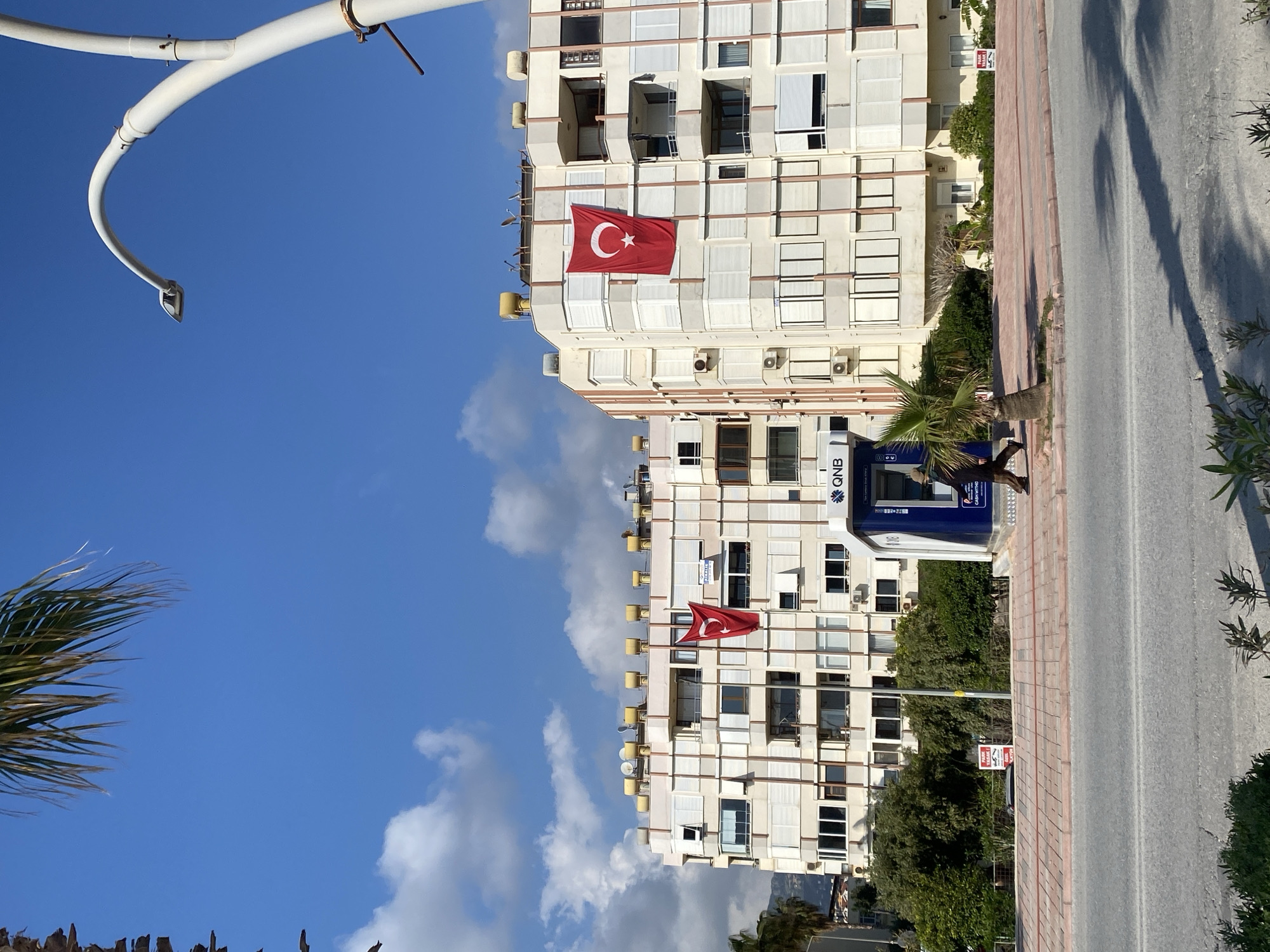 The image depicts a street scene in Alanya, Turkey. Two adjacent, multi-story apartment buildings with light-colored facades dominate the scene. Each building displays a large Turkish flag hanging from one of the upper floors. A QNB bank ATM is visible on the ground floor of one of the buildings. A narrow street runs alongside the building, partially obscured by vegetation. Palm trees are present in the foreground. A white, curved streetlight fixture is visible in the upper left corner of the frame. The sky is blue with scattered clouds. A person is using the ATM.