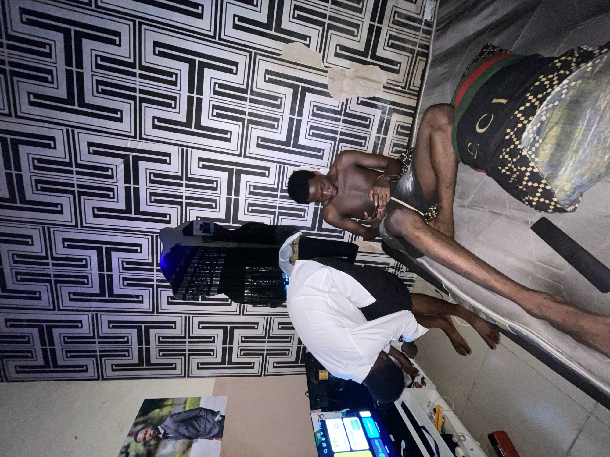 The image depicts the interior of a room, likely in a residence, with three individuals. A shirtless man is sitting on the floor near a wall with a geometric pattern, holding a mobile device. Another man, wearing a white shirt and dark pants, is also seated on the floor in front of a television screen displaying various icons. A third individual is positioned with their legs extended along the floor, wearing patterned shorts with a Gucci-branded waistband. The environment includes a television, wall art depicting a man in a suit, and a miscellaneous collection of objects on shelves below the television. There is no definitive visual evidence to confirm the location as Lokoja, Nigeria, or to specify any actions or interactions beyond the described scene.