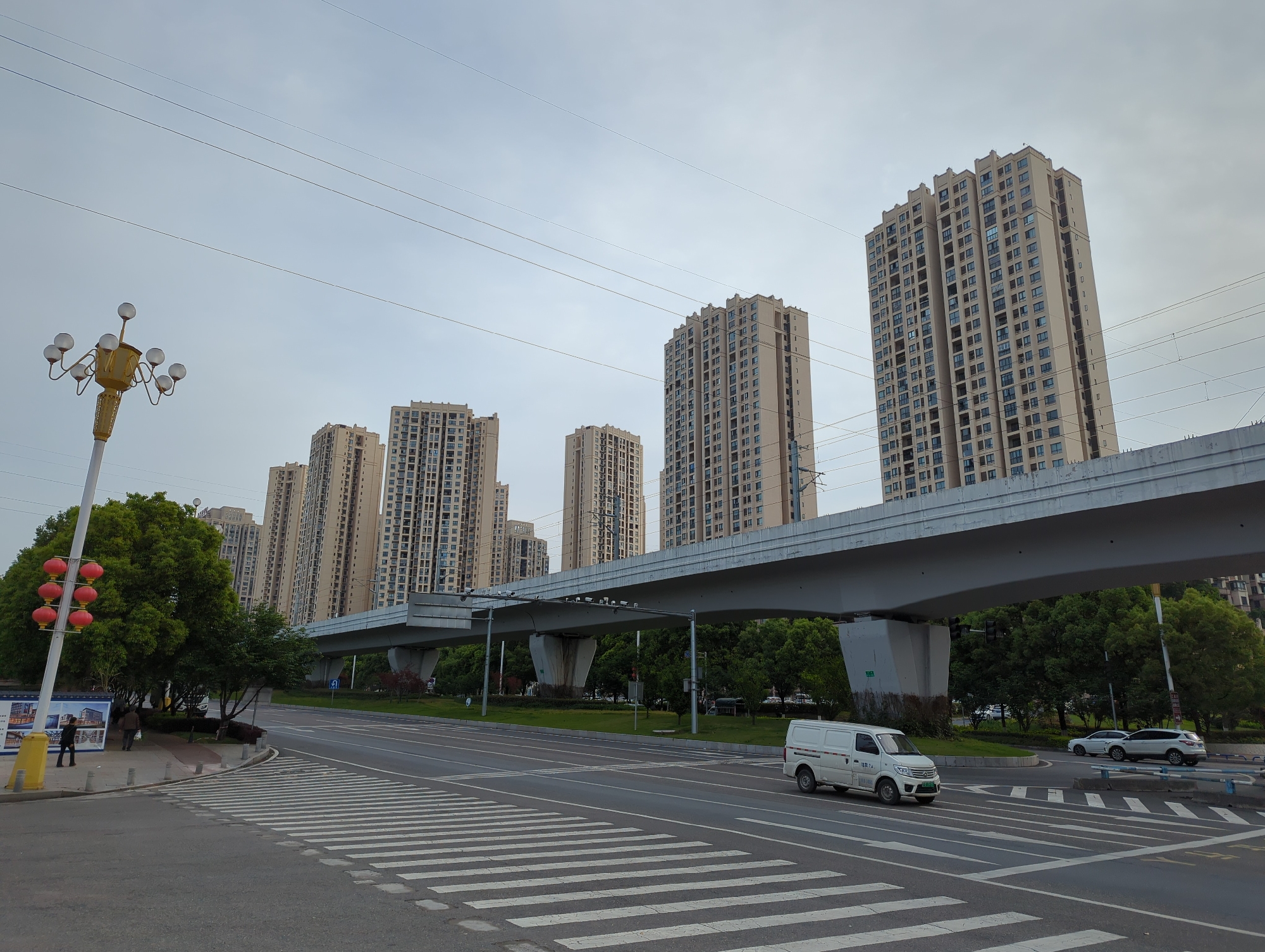 This wide-angle urban scene captures a street in Jiangjin, China, on an overcast day. The sky is a uniform light grey, with numerous overhead power lines traversing the frame.
The dominant subjects are several tall residential high-rise buildings with light-colored facades and many windows, extending into the background. An elevated concrete structure, likely for a metro or light rail, runs horizontally across the middle of the image, supported by thick pillars.
In the foreground, a multi-lane asphalt road features a large white zebra crossing. A white Changan brand delivery van is on the road, heading right, with Chinese characters "长安" (Cháng'ān) and what appears to be "货运专用" (freight dedicated) or "货运物流" (freight logistics) visible on its side. Further down the road, a white SUV is also present.
On the left side of the road, a sidewalk leads to a grassy area with trees. A tall, ornate golden street lamp with multiple spherical lights stands prominently. Nearby, another street lamp features a string of four red Chinese lanterns. Two pedestrians can be seen on the sidewalk next to a blue billboard displaying images of buildings and Chinese text, likely an advertisement for a property development. Green trees and bushes are interspersed along the road and under the elevated structure, providing natural elements. The scene depicts a modern, well-developed urban environment.