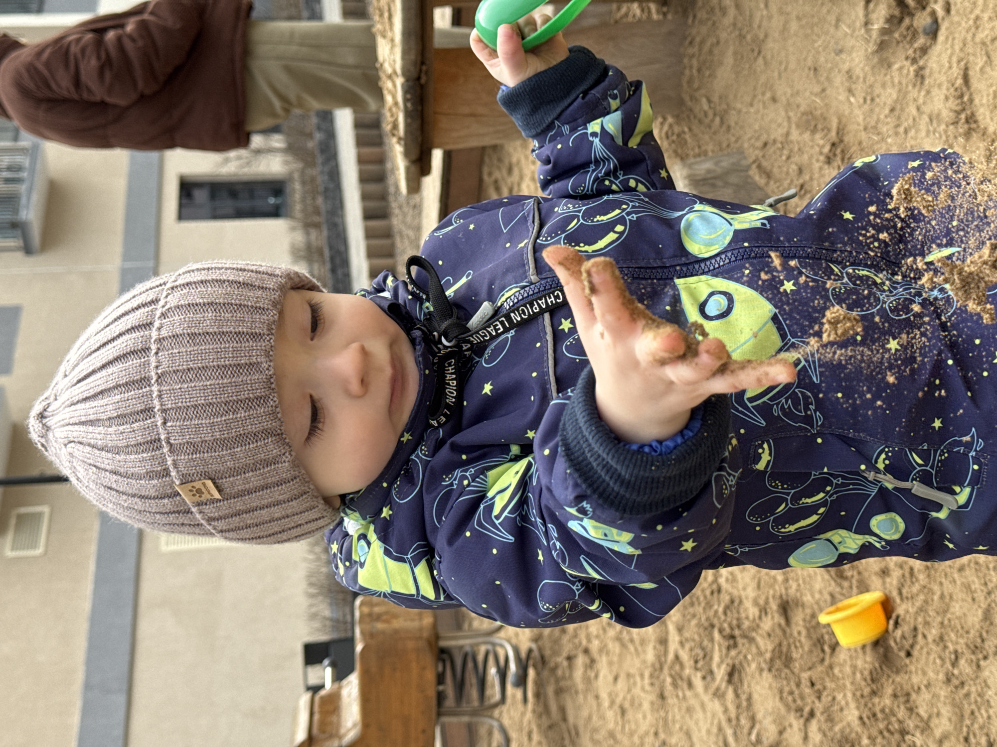 A child in a gray beanie and navy blue patterned snowsuit with lime green rockets is playing in a sandbox. The child's hands are covered in sand, and a small green plastic toy is held in their left hand. A yellow plastic toy is also visible in the sand. In the background, partially visible, is an adult figure wearing a brown jacket and beige pants. The scene appears to be a playground in a city, possibly Izhevsk, Russia, given the user's request. The environment suggests an outdoor setting with a sandy play area.