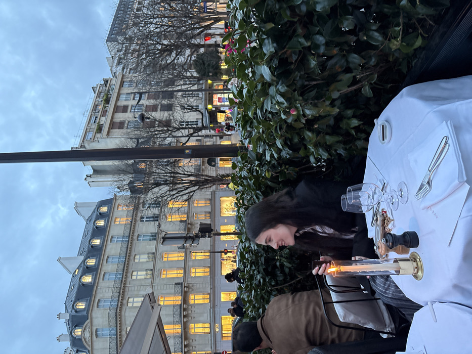 This photograph depicts an outdoor dining scene in Paris during dusk or early evening. A young woman with long, dark hair is seated at a table on the right, her head tilted downwards with a soft smile or amused expression as she looks at a lit candle. Another person, largely obscured and seen from behind, is seated across or next to her, wearing a brown jacket.
The table is covered with a crisp white tablecloth and set with two empty wine glasses, a salt and pepper grinder, and cutlery placed on a white napkin. A prominent feature is the tall, cylindrical candle holder with a glowing candle, casting a warm light on the immediate area.
In the background, characteristic Parisian buildings with intricate facades and dormer windows line the street. Many of their windows are illuminated with a warm yellow light, standing out against the overcast, muted blue sky. Bare trees, indicative of late autumn or winter, are scattered along the street. Streetlights and traffic signals are also visible further down. The overall ambiance is cozy and urban.
On the white napkin under the cutlery, the faint partial text "AVEN" can be seen.