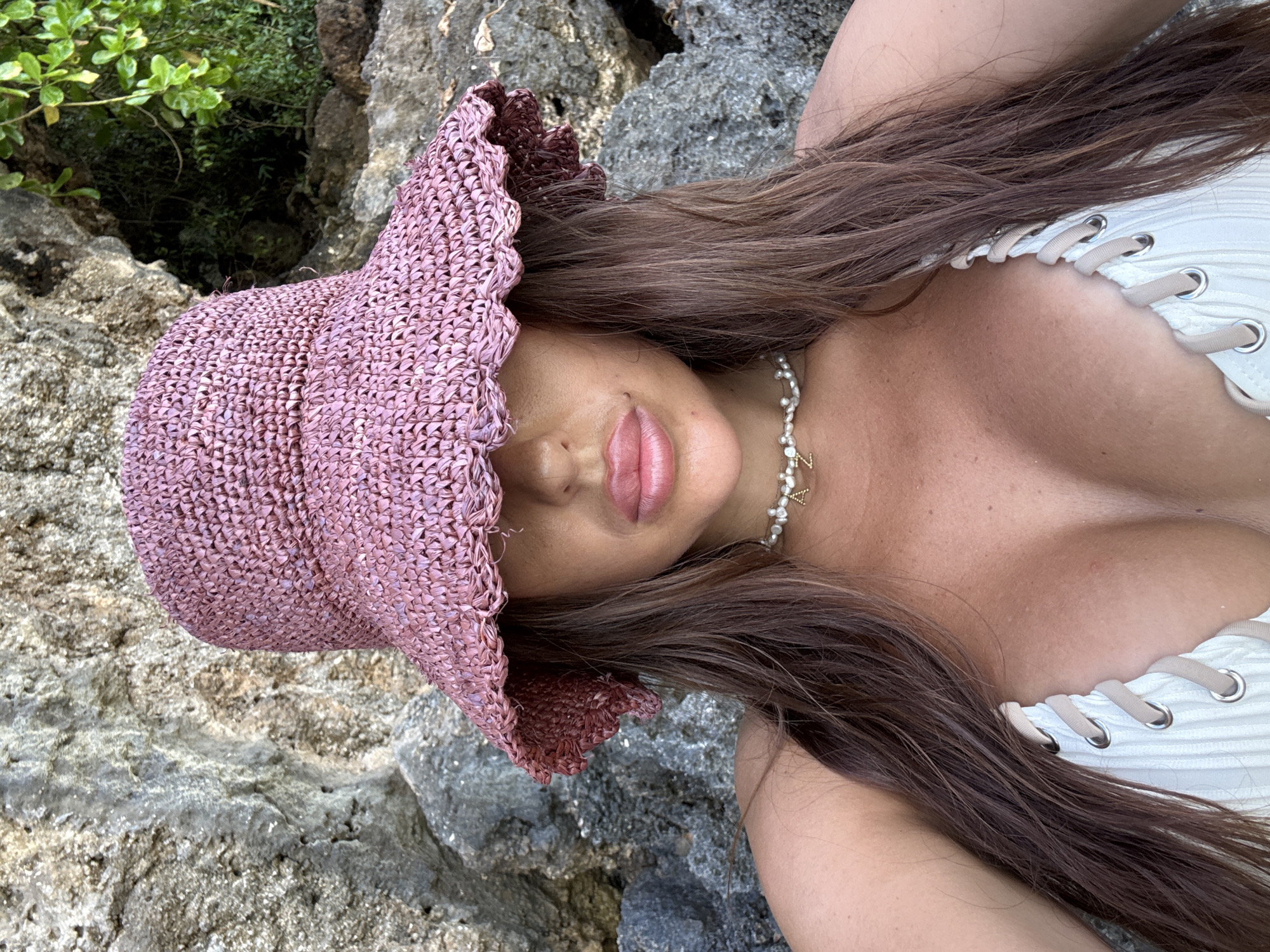A woman lies on a rocky surface, possibly near a beach, in Kuta Selatan, Indonesia. She is wearing a white top with a laced front and a light-purple, floppy hat. Her long brown hair lies on the rock, next to her. A pearl necklace with a gold letter “Z” pendant is around her neck. The rocky surface is near some greenery.