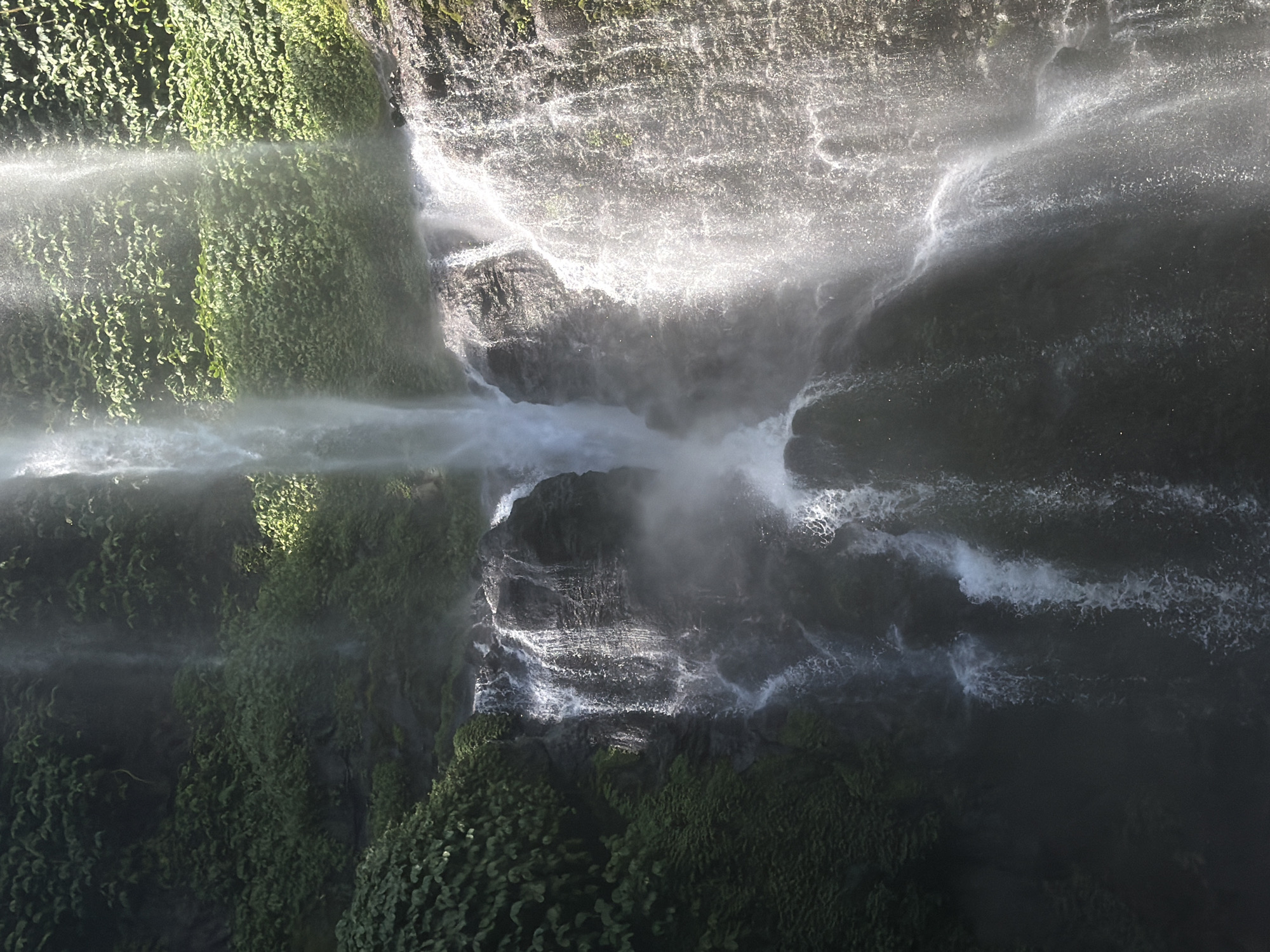 A tall, narrow waterfall cascades down a rocky cliff face covered in lush green foliage. The water is bright white and appears to be illuminated by sunlight. The surrounding rocks are dark and textured, with some areas appearing wet and mossy. The vegetation is dense and a deep green, with various types of leaves visible. The water creates a mist as it falls, which softens the appearance of the rocks and vegetation. The overall impression is one of a powerful and natural scene. The waterfall is located in the city of Sawan, Indonesia.