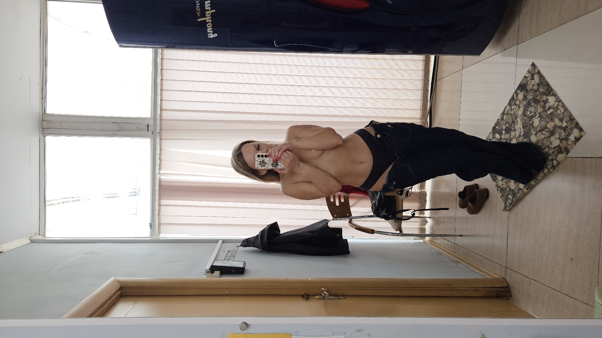 The image depicts a room with a partially visible window on the left and window blinds behind a person taking a selfie. The person is fair-skinned, wearing dark-colored underwear and jeans, and holding a mobile phone. To their right, on the floor, are a pair of brown shoes and a patterned rug. A jacket and other items are positioned on a surface. The location in the city of Sevastopol, Ukraine, cannot be verified.