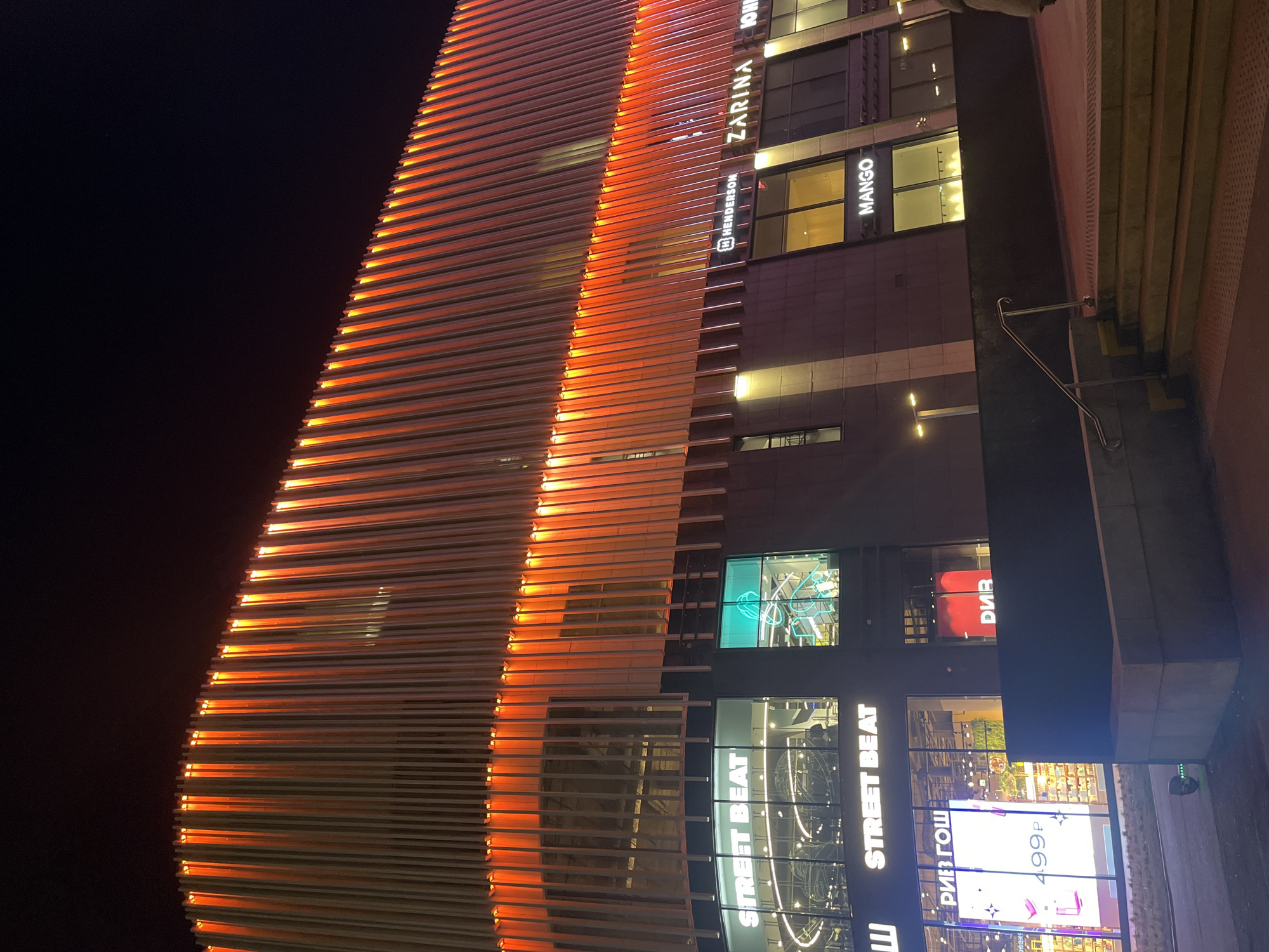 The image depicts the facade of a building at night, likely located in Saint Petersburg, Russia. The architectural style is modern with a prominent feature being horizontal slats that are illuminated with an orange glow. The building has commercial spaces, recognizable by store names like "ZARINA," "HENDERSON," "MANGO," and "STREET BEAT." Large display windows showcasing retail goods are also visible. The sky is dark, indicating nighttime, and the surrounding area is partially visible, suggesting an urban setting with pedestrian access.