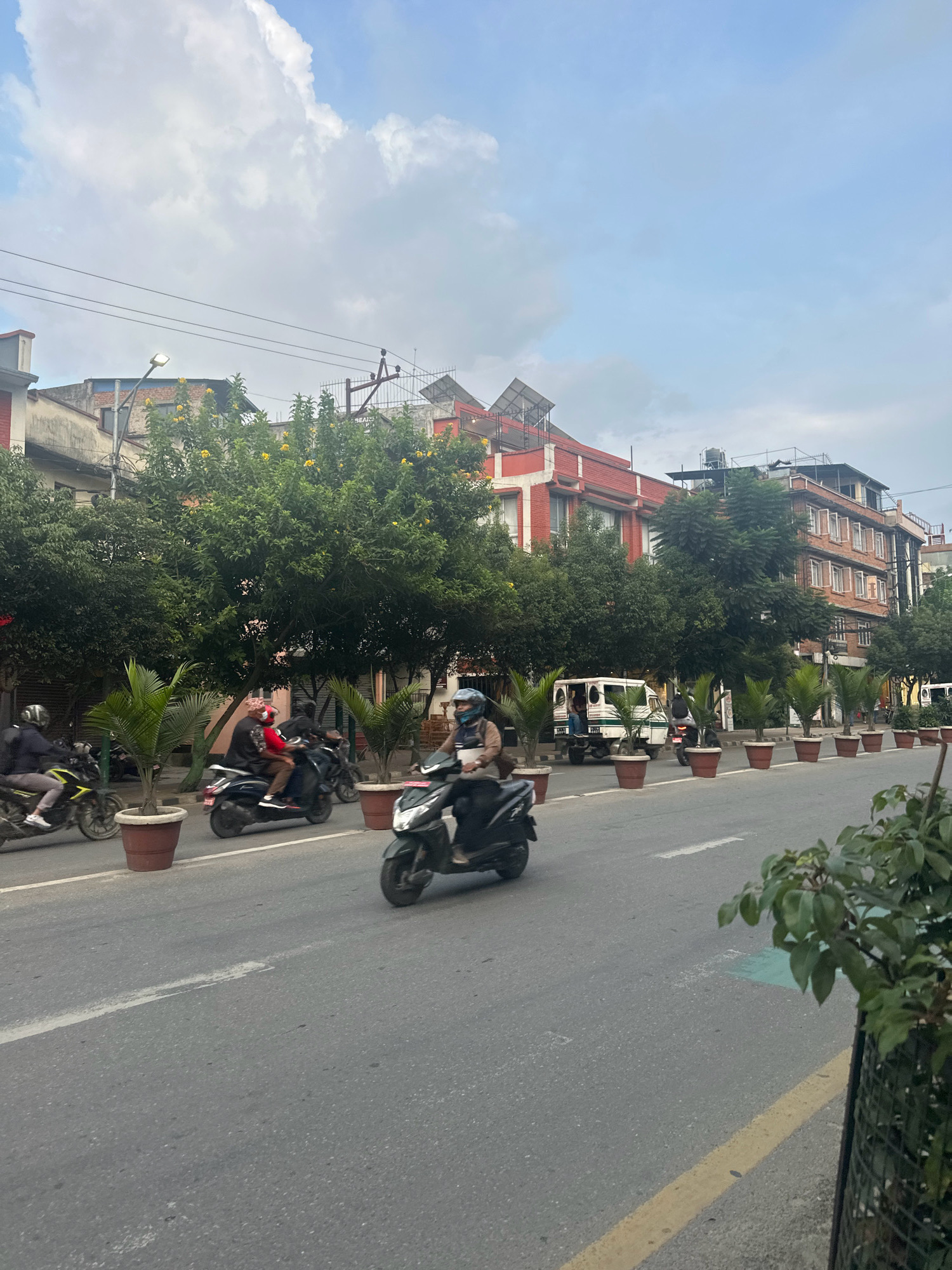 A street scene in Lalitpur, Nepal, captures a moderately busy road with several motorcycles and a van. The road is paved with asphalt and marked with white lines. On the left side of the road, trees in large brown planters line the pavement. Behind the trees, multi-story buildings with red and beige facades are visible. On the right side, a row of similar planters with palm trees separates the road from the buildings. The sky above is partly cloudy with a mix of white clouds and blue sky. Several individuals are riding motorcycles, some with passengers, moving along the road. A white van is also present on the street. The overall impression is of an urban environment on a daytime.