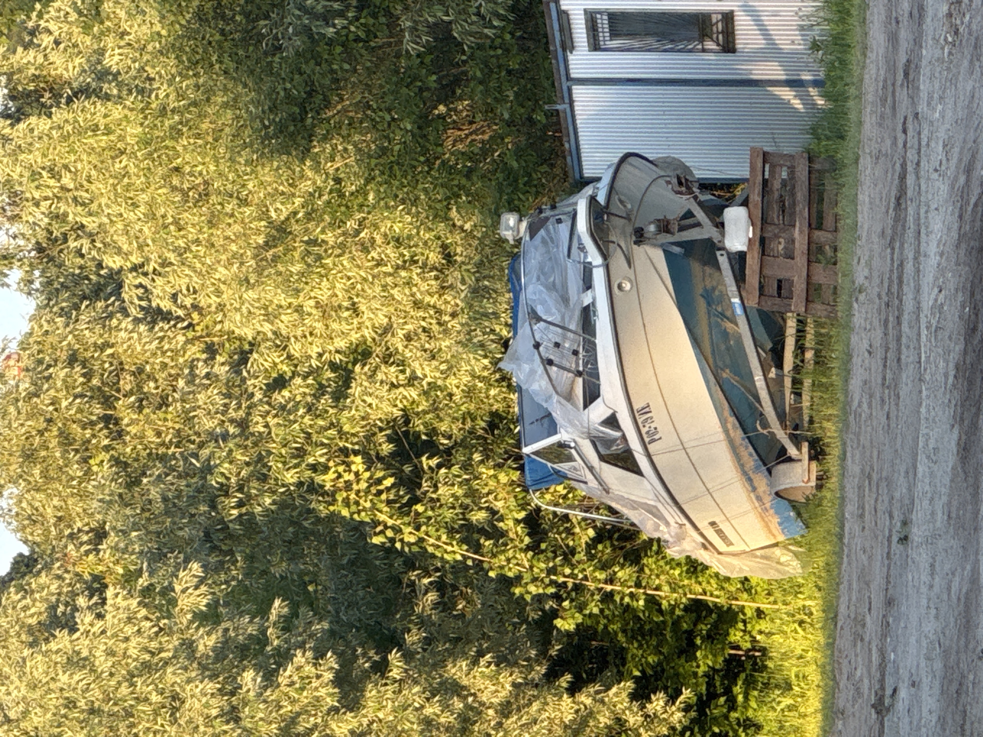 A boat is propped up by a wooden pallet outside of a metal building, partially obscured by foliage. The location is unknown, and the city is unknown.