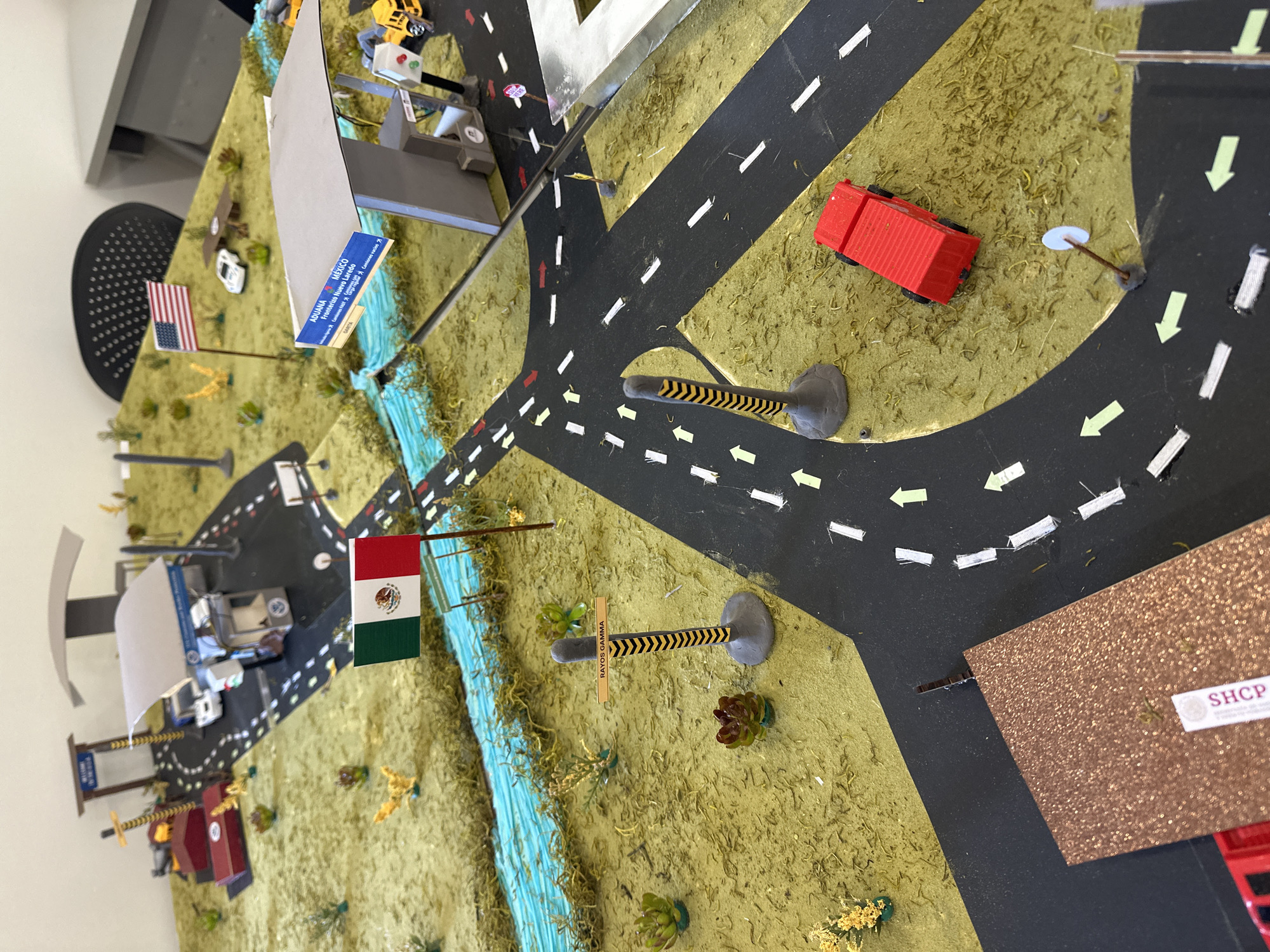 This is a model of the US-Mexico border. A blue painted area represents the river that divides the two countries. The United States is on the top side of the image, denoted by the American flag, road signs in English, and a US Customs and Border Patrol checkpoint. Mexico is on the bottom half of the image, indicated by the Mexican flag and "RAYOS GAMMA" road signs. There are roads with white and green directional arrows in both countries and small vehicles scattered throughout the model. The building labeled "SHCP" is visible on the Mexican side of the border. This model is not located within San Bernardino Tlaxcalancingo, Mexico. Instead, it is representative of the border between the United States and Mexico generally, and not a specific geographic location.