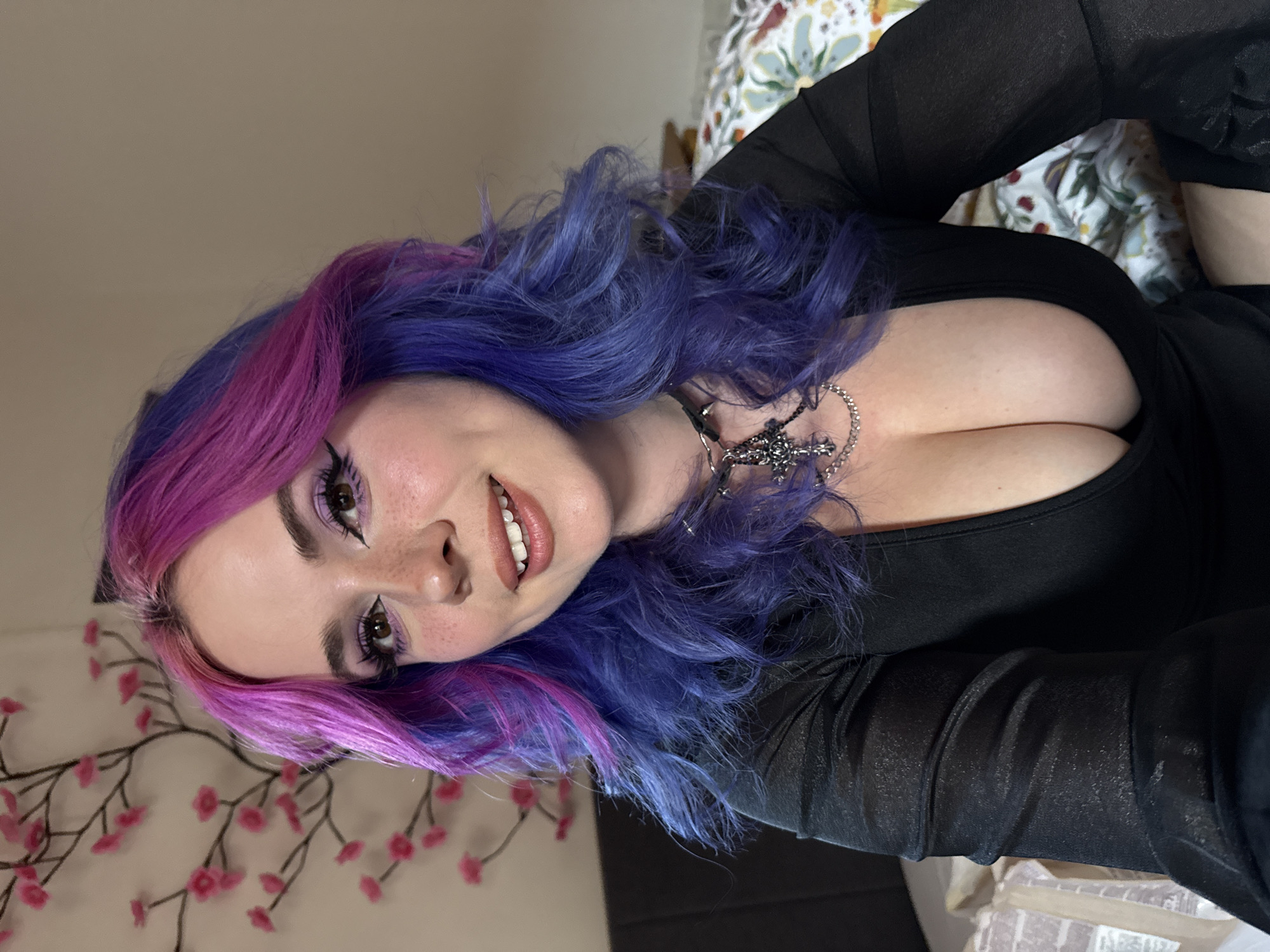 A young woman is captured in a close-up, medium shot, appearing to be lying down or leaning back, looking towards the camera with a slight smile. The setting is indoors, possibly a bedroom or living area in the City of Zagreb, Croatia. She has striking, long, wavy hair, predominantly purple with prominent pink sections framing her face. Her makeup includes detailed eyeliner, purple eyeshadow, mascara, and a subtle lip color, complemented by visible freckles across her nose and cheeks.
She is wearing a black, low-cut top with a scoop neck, featuring long, sheer, textured sleeves. Around her neck is a silver-toned chain necklace with an intricate, dark, cross-shaped pendant. To the left of the frame, decorative string lights with pink, cherry blossom-like flowers are visible against a light-colored wall. The background on the right features a patterned fabric, likely part of bedding or a cushion, with a floral design in muted colors. The lighting is soft and even, highlighting her features and hair. No text is visible in the image. Her expression suggests a pleasant or contented mood while posing for the photograph.