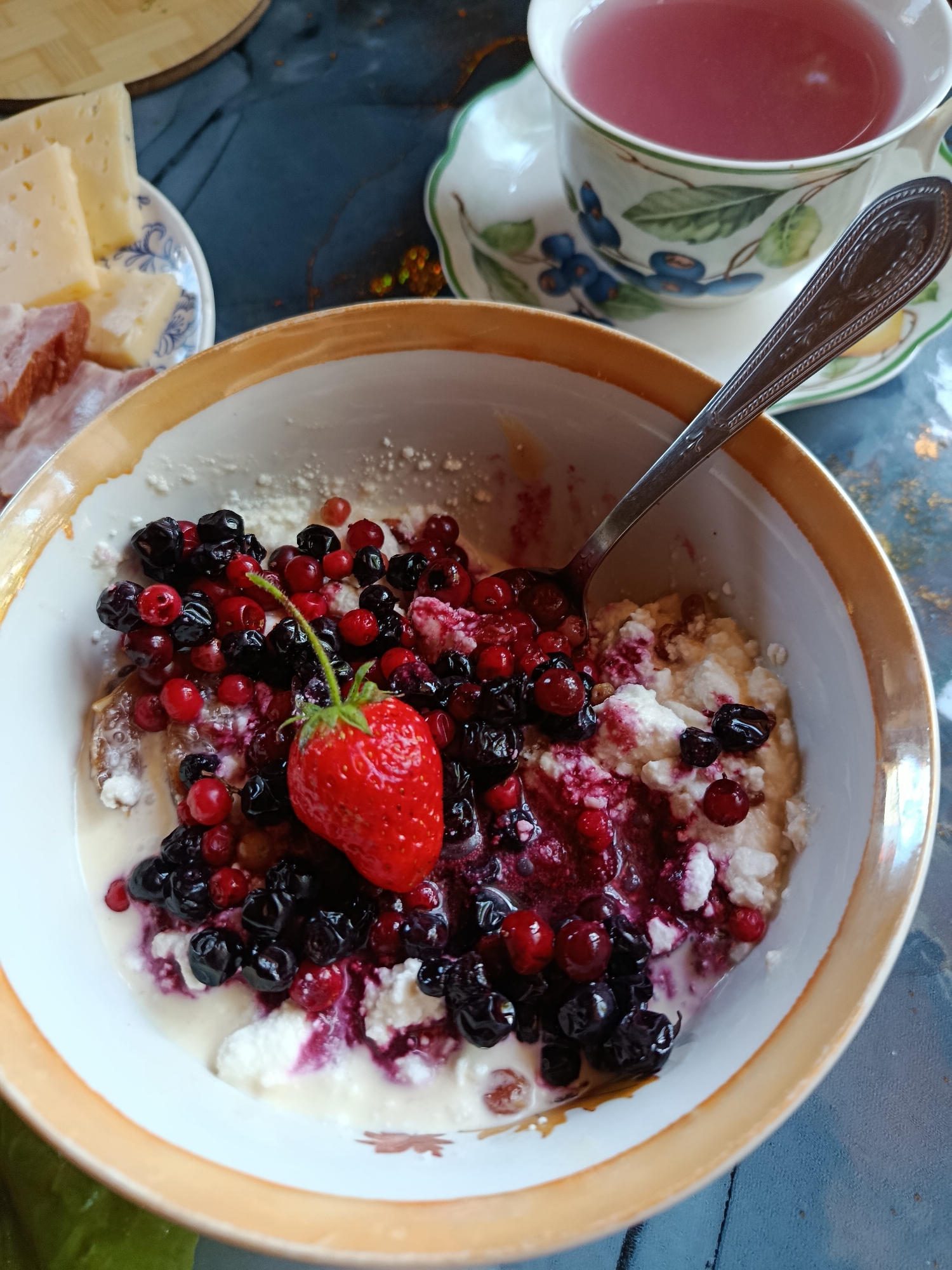 This high-angle shot captures a casual meal spread across a dark blue or gray patterned table. The primary subject is a white bowl with a golden-orange rim, containing what appears to be cottage cheese or a similar dairy product. It is lavishly topped with a colorful medley of fresh berries, including dark blue blueberries, bright red redcurrants, and a prominent whole red strawberry with its green stem. A silver spoon, featuring an ornate handle, rests within the bowl, partially submerged in the berries and a creamy liquid that has mixed with the berry juices, creating streaks of purple and red.
In the upper right background, a decorative teacup and saucer are visible. The teacup, adorned with a pattern of blue berries and green leaves, holds a reddish-pink beverage, likely berry tea. To the upper left background, a small white plate with blue accents contains several slices of light yellow cheese and what appears to be cured meat, possibly bacon or ham. A partial view of a wooden cutting board can also be seen behind this plate.
The scene suggests a daytime meal, possibly breakfast or brunch, enjoyed indoors. The fresh ingredients and wholesome presentation evoke a sense of comfort and pleasantness. No people are visible, and there is no discernible text within the image. The arrangement implies a leisurely and satisfying eating experience in a home environment.