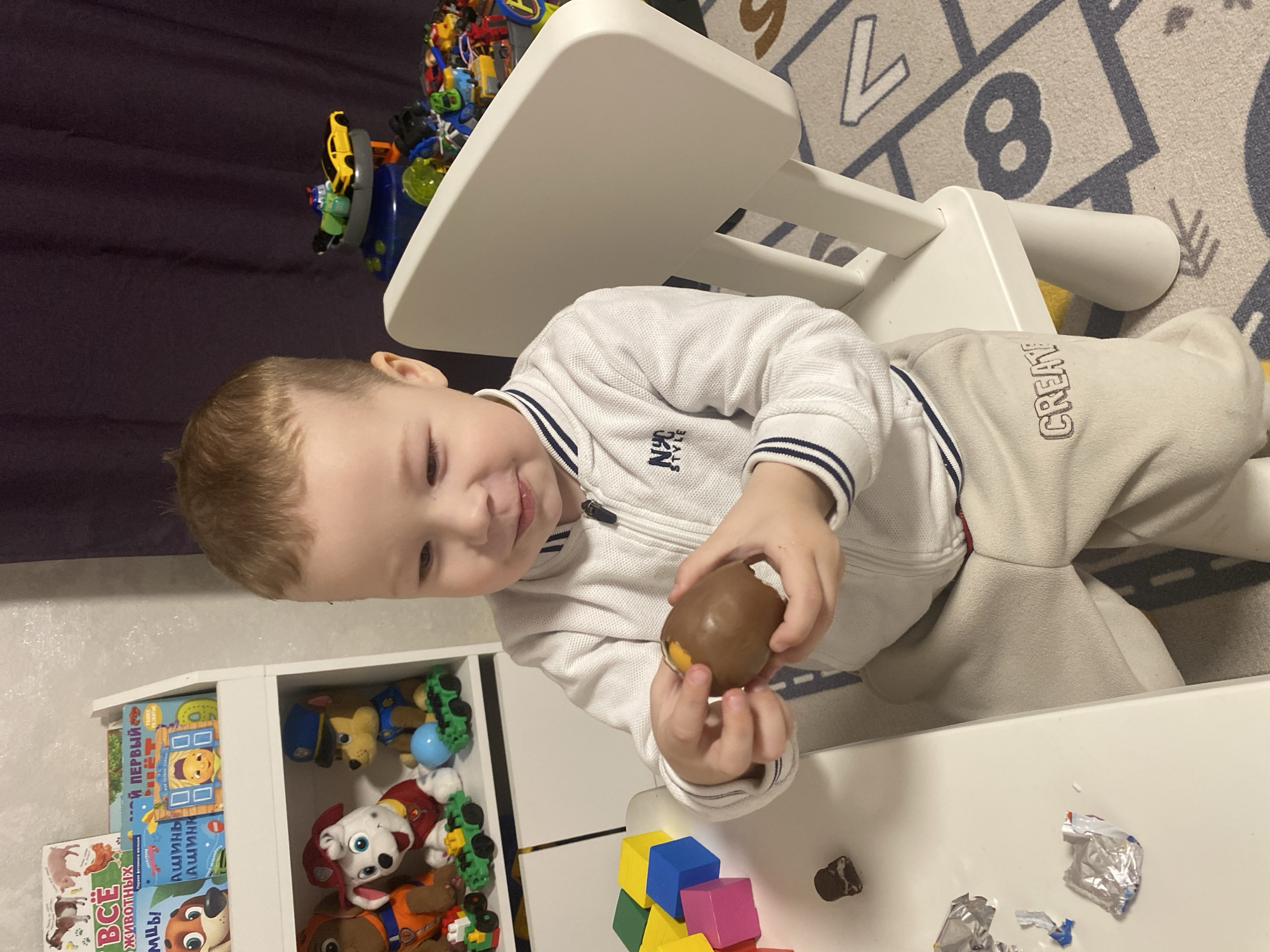 A young child is sitting at a white table. He is wearing a white zip-up jacket and light-colored pants with the word "CREATE" on the leg. He is holding a chocolate egg in his hands and appears to be opening it. On the table are colorful building blocks and pieces of foil wrapping. Behind him is a white storage unit filled with toys and books. In the background, there is a rug with numbers and a curtain. There is no indication of the location or that it is in the city of Sochi, Russia.