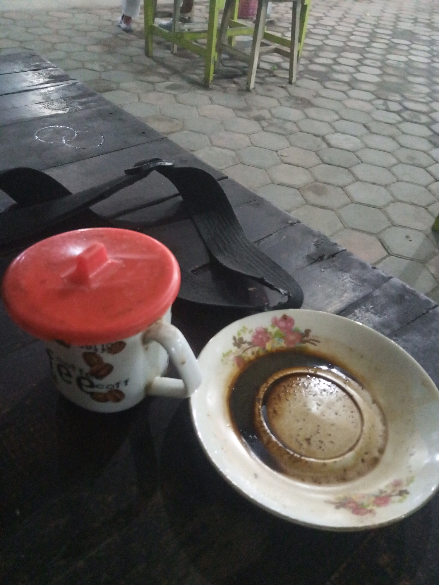 A close-up, low-angle, outdoor shot shows a table surface and a tiled ground in the city of Gayaman, Indonesia. On the table, a white mug with a red lid sits to the left, and a saucer with dark liquid is on the right. The mug has brown coffee beans and the word "coffee" printed on it. A black sandal is positioned behind the mug, partially covering the table. The tiled ground in the background consists of hexagonal pavers. In the upper background, a portion of a green table and legs of chairs is visible, with two legs of chairs and two pairs of legs with white pants and dark shoes standing on the tiled ground.