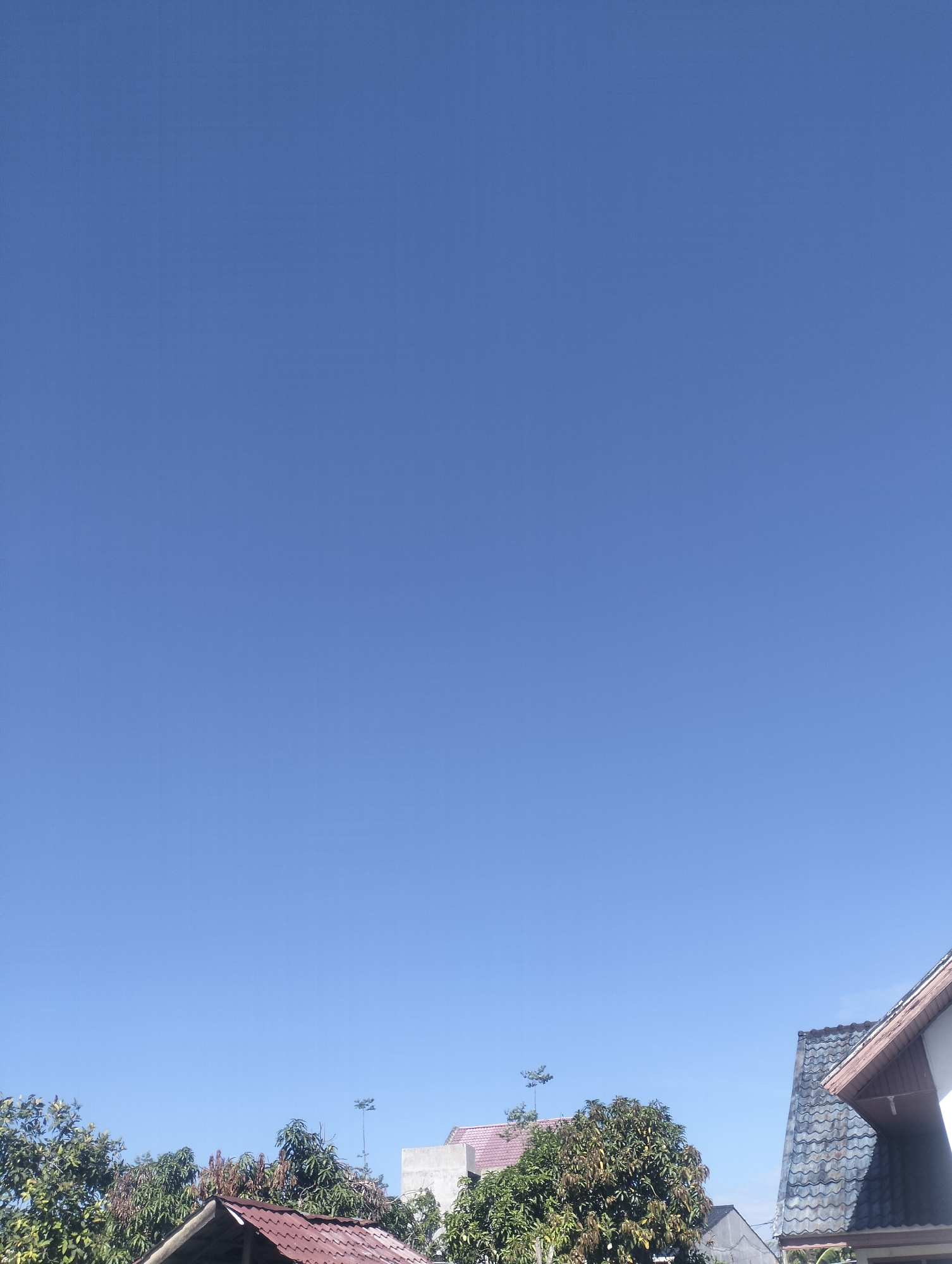 The image shows a view of a clear blue sky above a residential area in Lhokseumawe, Indonesia. Several rooftops are visible, including a dark gray tiled roof, a red tiled roof, and sections of other buildings interspersed with trees. There are two objects resembling antenna towers in the far background.