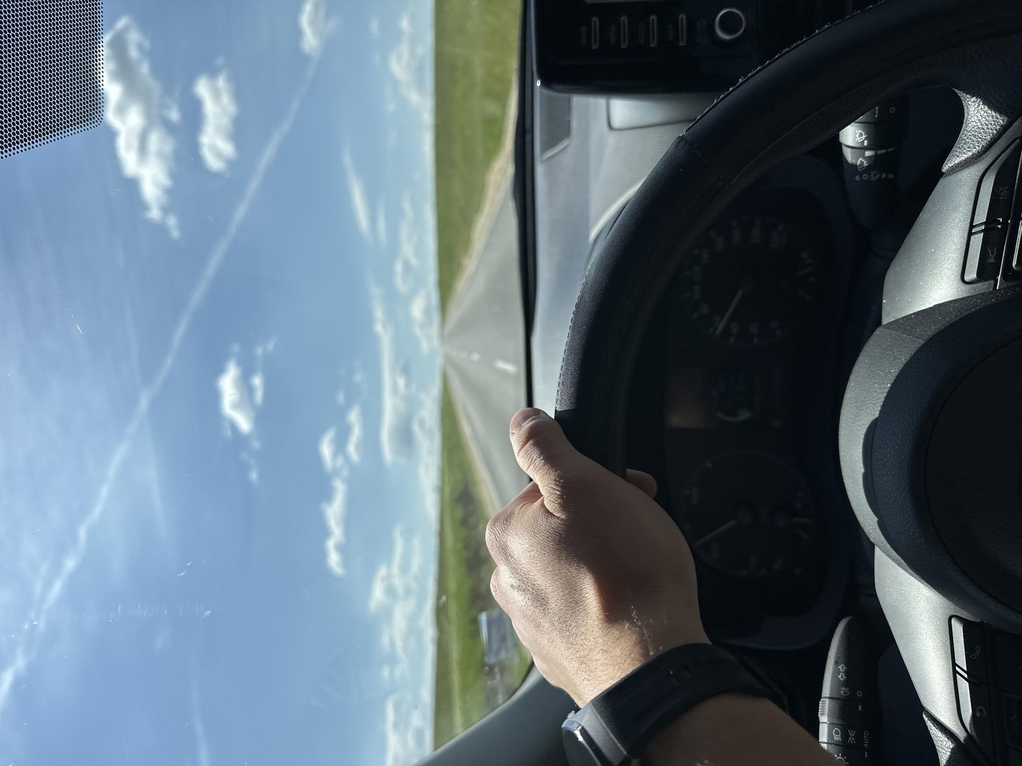 A person is driving a car on a road, possibly in the countryside, as seen through the windshield. The sky is blue with scattered clouds. The driver's hand is visible on the steering wheel. Part of the car's dashboard and instrument panel are also in the frame. There is no information in the image that suggests that the location is Lukovit, Bulgaria.