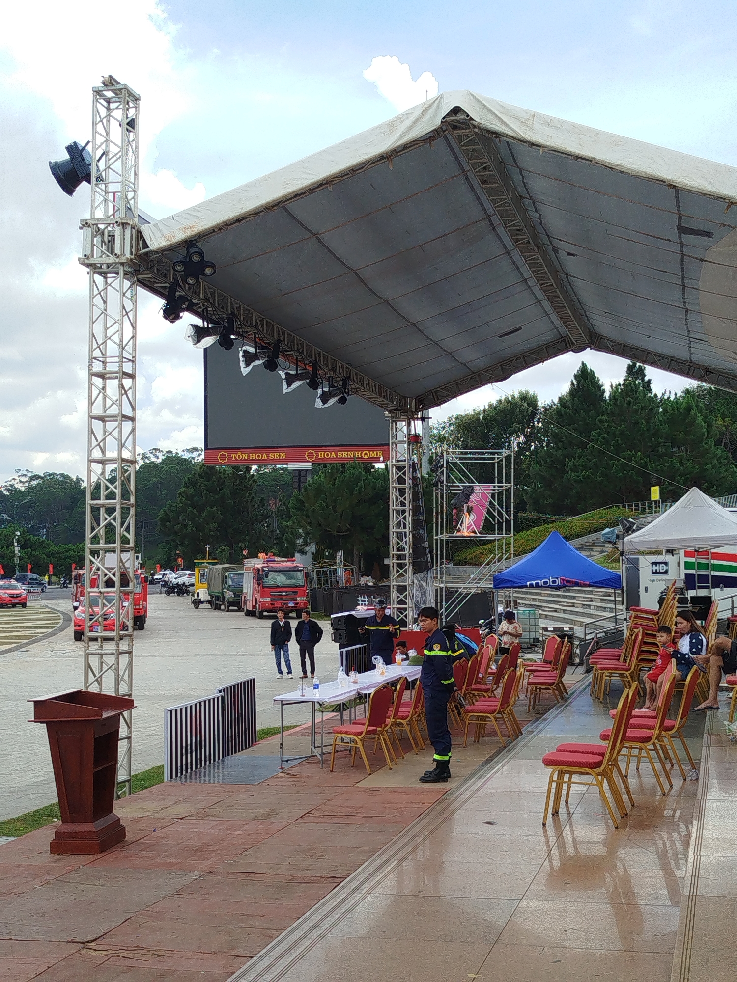 This image depicts an outdoor event setup in Da Lat, Vietnam, likely at a venue with tiered seating and a large stage area. A substantial stage structure, covered by a white tarp, is prominent, with scaffolding and lighting rigs visible. In front of the stage, a series of red chairs are arranged, some occupied by attendees. A dark wooden podium stands to the left, and a long table with chairs is set up further back. Several red trucks, resembling fire trucks, are parked on a paved area behind the stage, suggesting a public event or ceremony. The background shows lush green trees and a cloudy sky. Several individuals, some in uniform, are present, overseeing the setup and possibly attending the event.