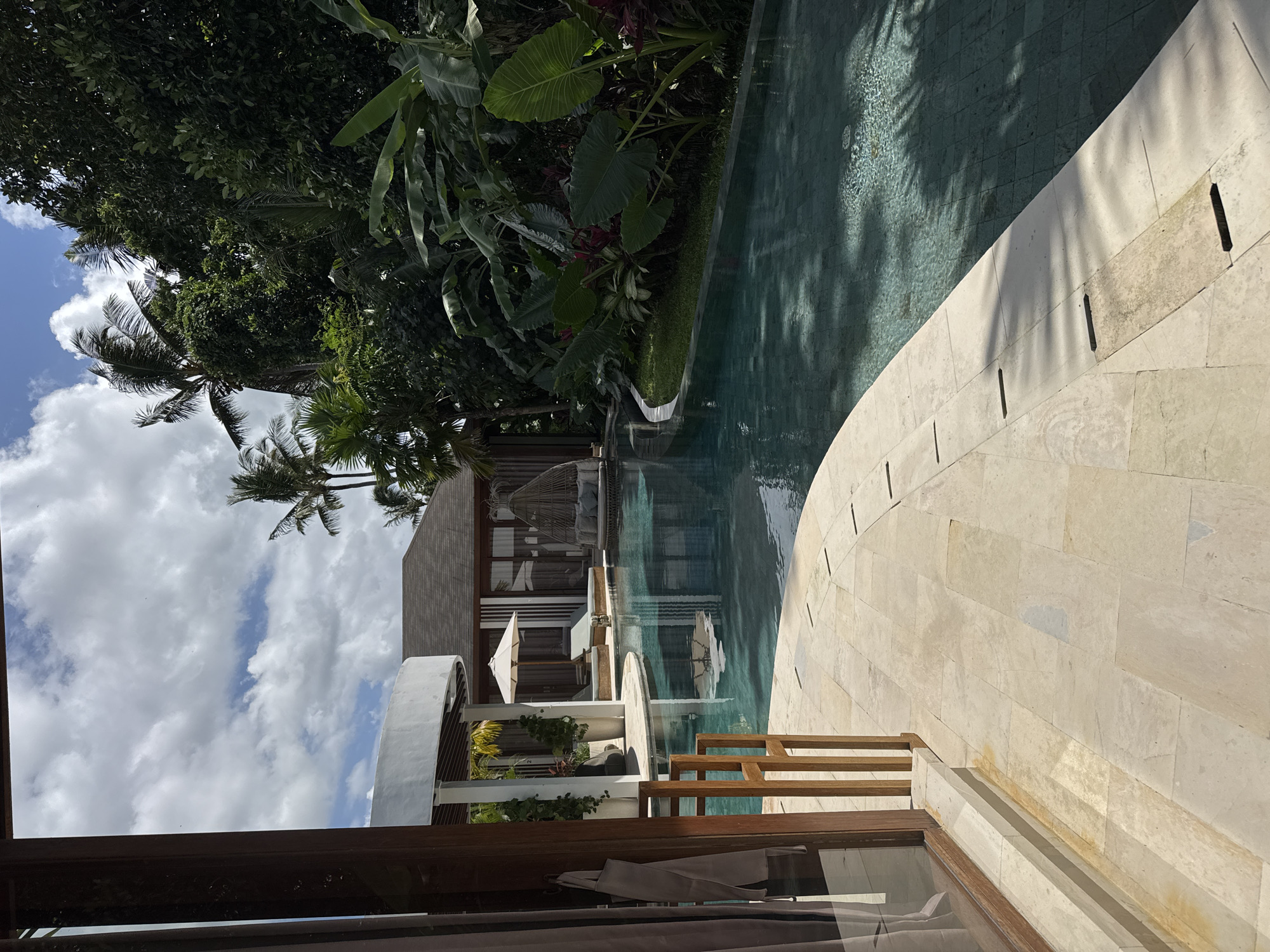The image depicts a tropical resort in the Ubud District of Indonesia. A long, narrow, curvilinear swimming pool occupies a significant portion of the scene, reflecting the surrounding lush vegetation and structures. The pool is bordered by a light-colored tiled walkway. Beyond the pool, a modern multi-story building is visible, featuring white architectural elements and light-colored cladding. Parasols, outdoor furniture, and verdant plants adorn the outdoor spaces associated with the building. The sky is visible in the upper-left portion of the frame, revealing scattered clouds and a few palm trees. A wooden frame from a window or doorway is visible in the lower foreground, providing a framed view of the exterior scene.