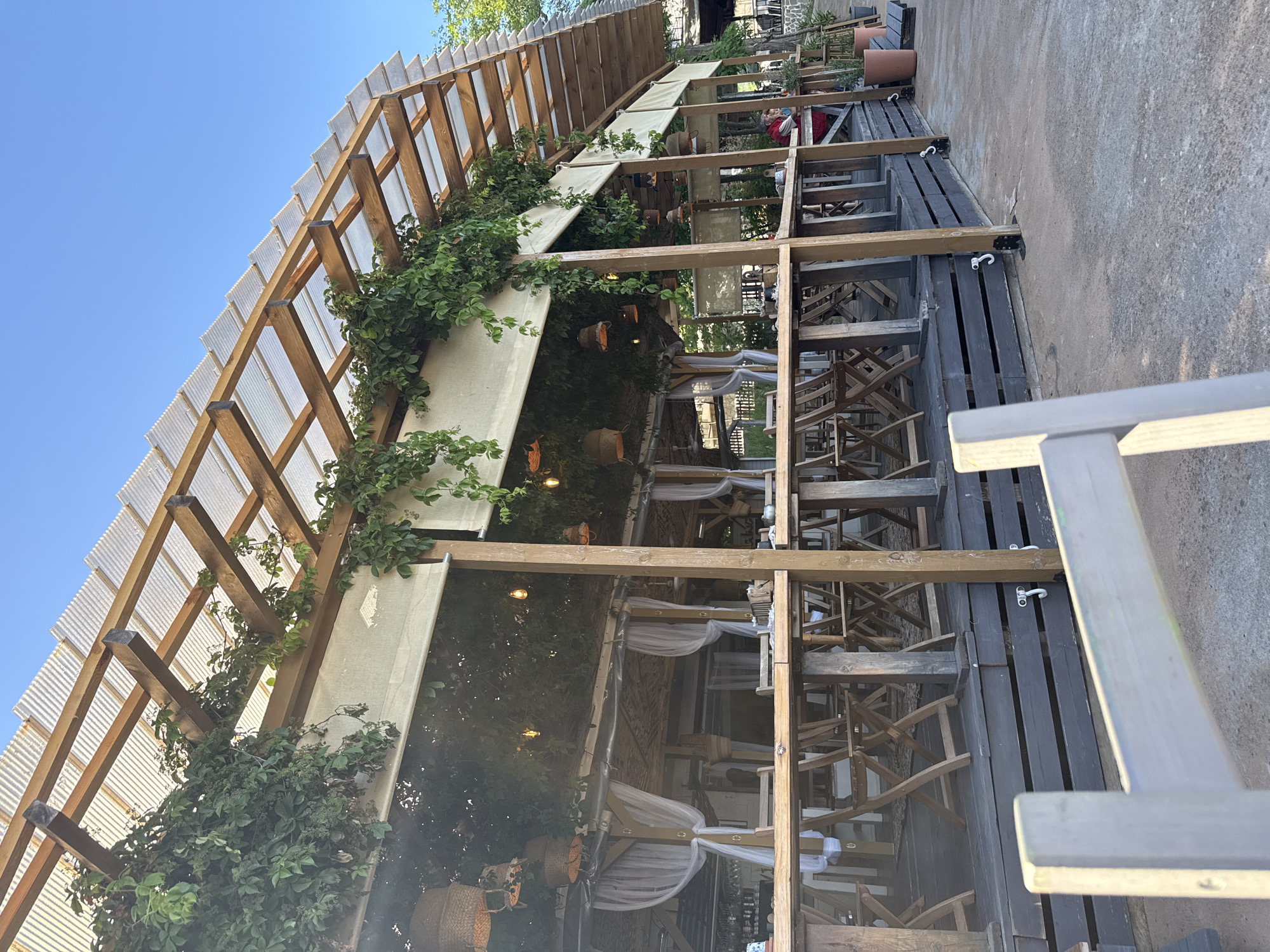 The image depicts an outdoor dining area with a wooden structure covered by a corrugated plastic roof. The structure is adorned with green vines and hanging baskets. White curtains are draped along the sides, creating a relaxed, open-air ambiance. Numerous wooden tables and chairs are arranged on a dark wooden deck, suggesting a restaurant or cafe setting. The foreground shows a light-colored wooden railing and a concrete ground surface. In the background, there are glimpses of more seating, potted plants, and a clear blue sky. The overall scene suggests a sunny day at an establishment located in Tsarevo, Bulgaria.