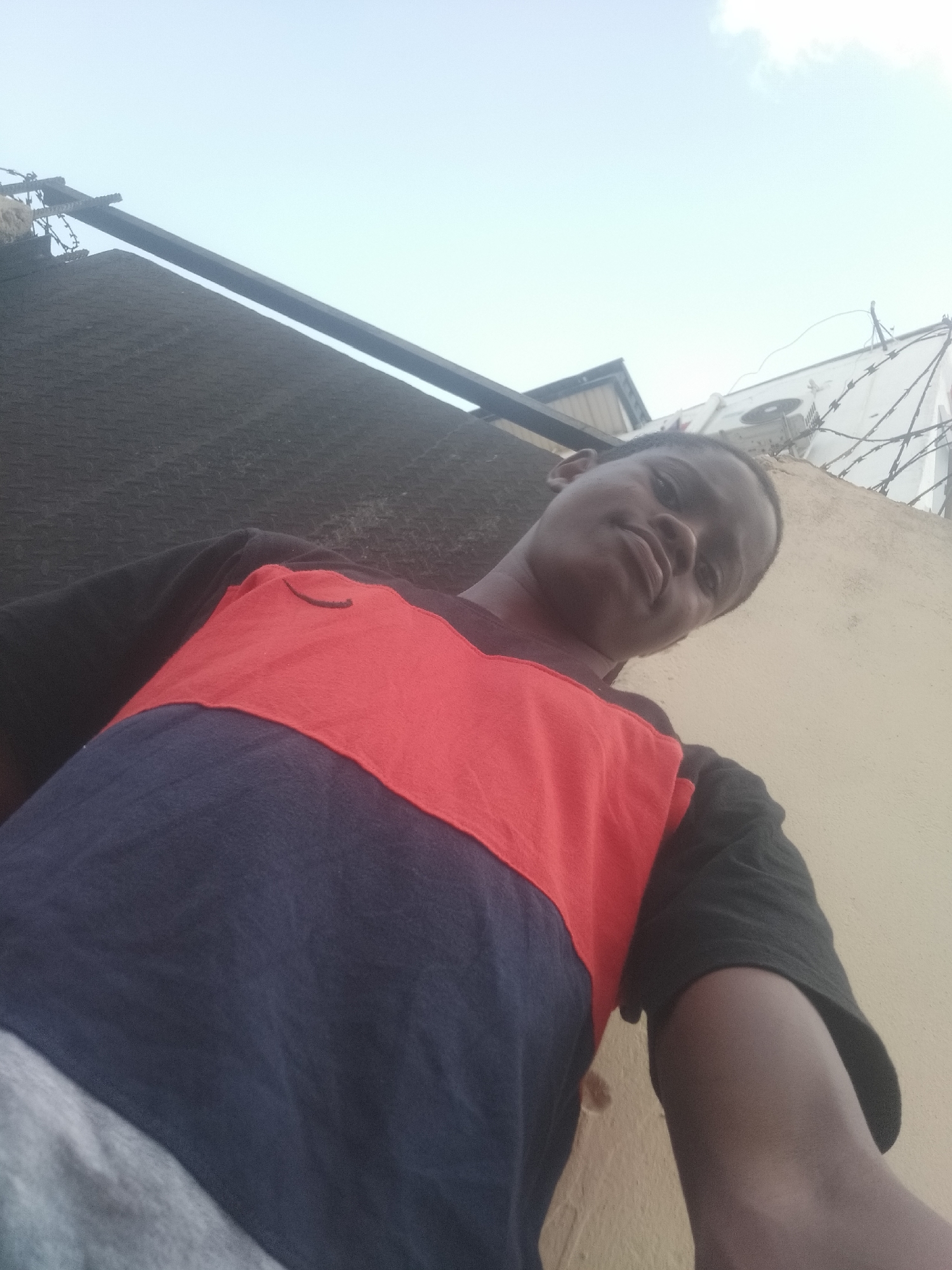 A low-angle, outdoor selfie taken in Shangisha, Nigeria, depicts a dark-skinned male wearing a short-sleeved t-shirt with a red and blue color block design over a gray bottom layer. The sleeves and collar of the t-shirt are black. The man is positioned in the lower half of the frame. Above him is a cloudy, pale blue sky. Behind and to the left of the man, there is a section of black corrugated metal fencing with a metal support structure at the top. To the right and behind the man, there is a beige wall with barbed wire along the top. An air conditioning unit is also visible near the top right of the frame.