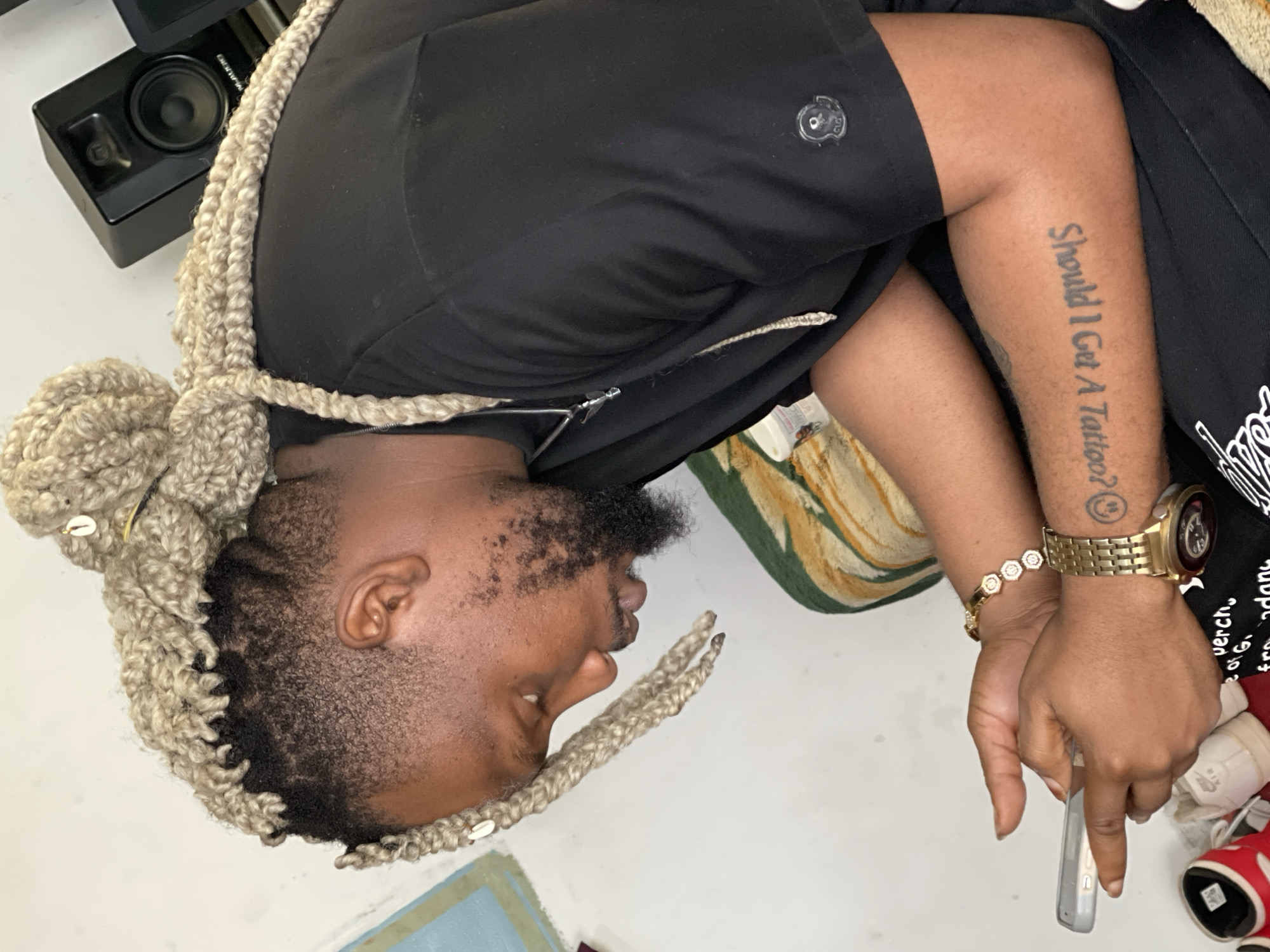 A side profile of a Black man with light-colored dreadlocks, a beard, and a black t-shirt is shown. He is seated and looking down at a smartphone he is holding in his hands. He is wearing a gold bracelet on his left wrist and a gold watch on his right wrist. A tattoo is visible on his right forearm. The background is a plain wall with some decor. The man appears to be indoors, in a residential setting. The overall lighting is bright and natural.