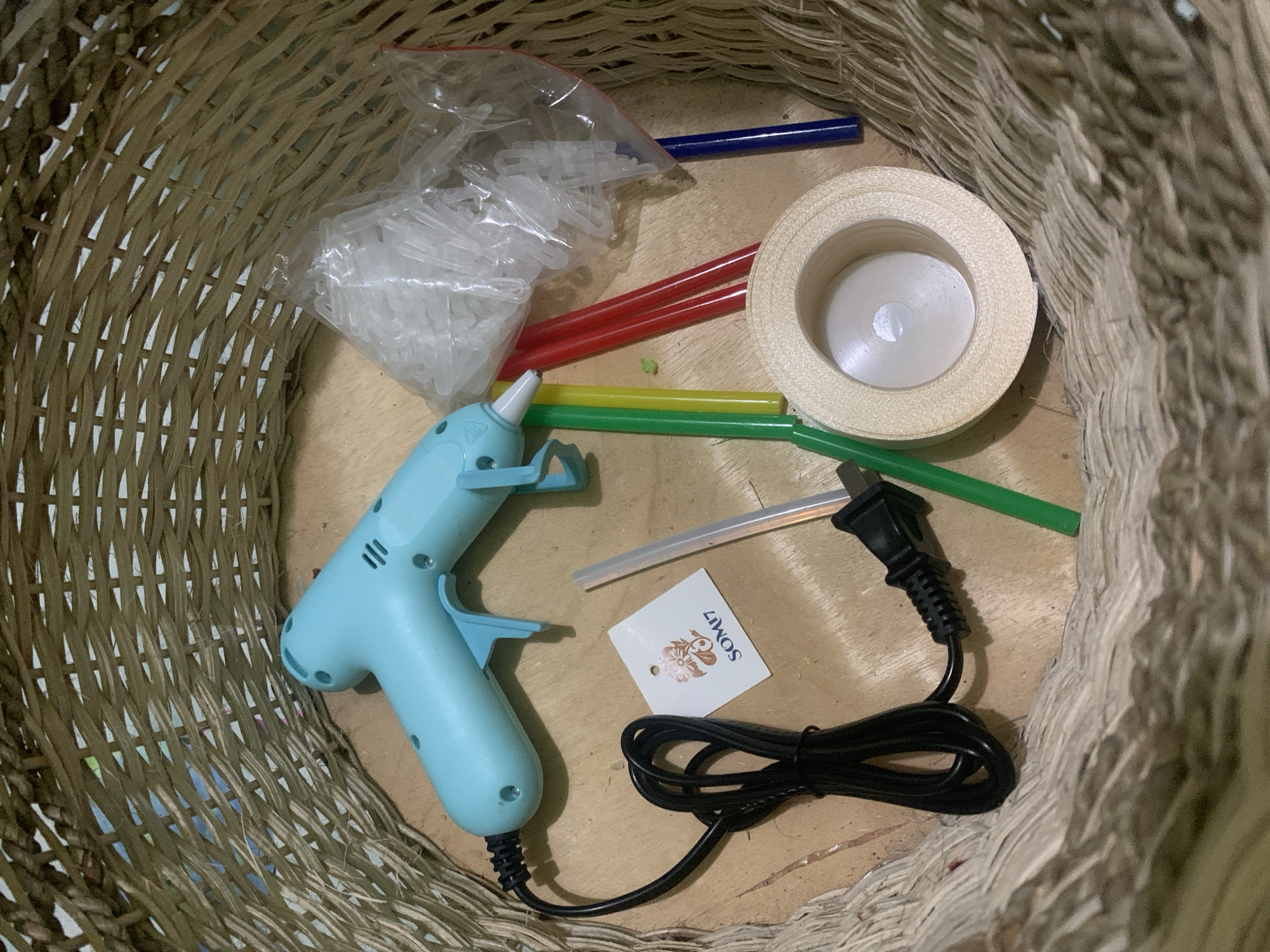 Inside a woven basket, a light blue hot glue gun is placed on a wooden surface. Beside the glue gun, a small white tag with a floral design and the text "SOM17" is visible. Several colorful glue sticks, including green, yellow, red, and blue, are arranged next to the gun. A clear plastic bag filled with white glue sticks is also present. In the foreground, a roll of light beige ribbon lies on the surface. The basket appears to be made of natural fibers and is woven in a crisscross pattern. The scene is illuminated by overhead lighting, casting subtle shadows. The location is within the city of Kuala Pembuang, Indonesia, specifically on a table or flat surface inside a dwelling. There are no people or actions depicted in the image; it is a static arrangement of craft supplies.
