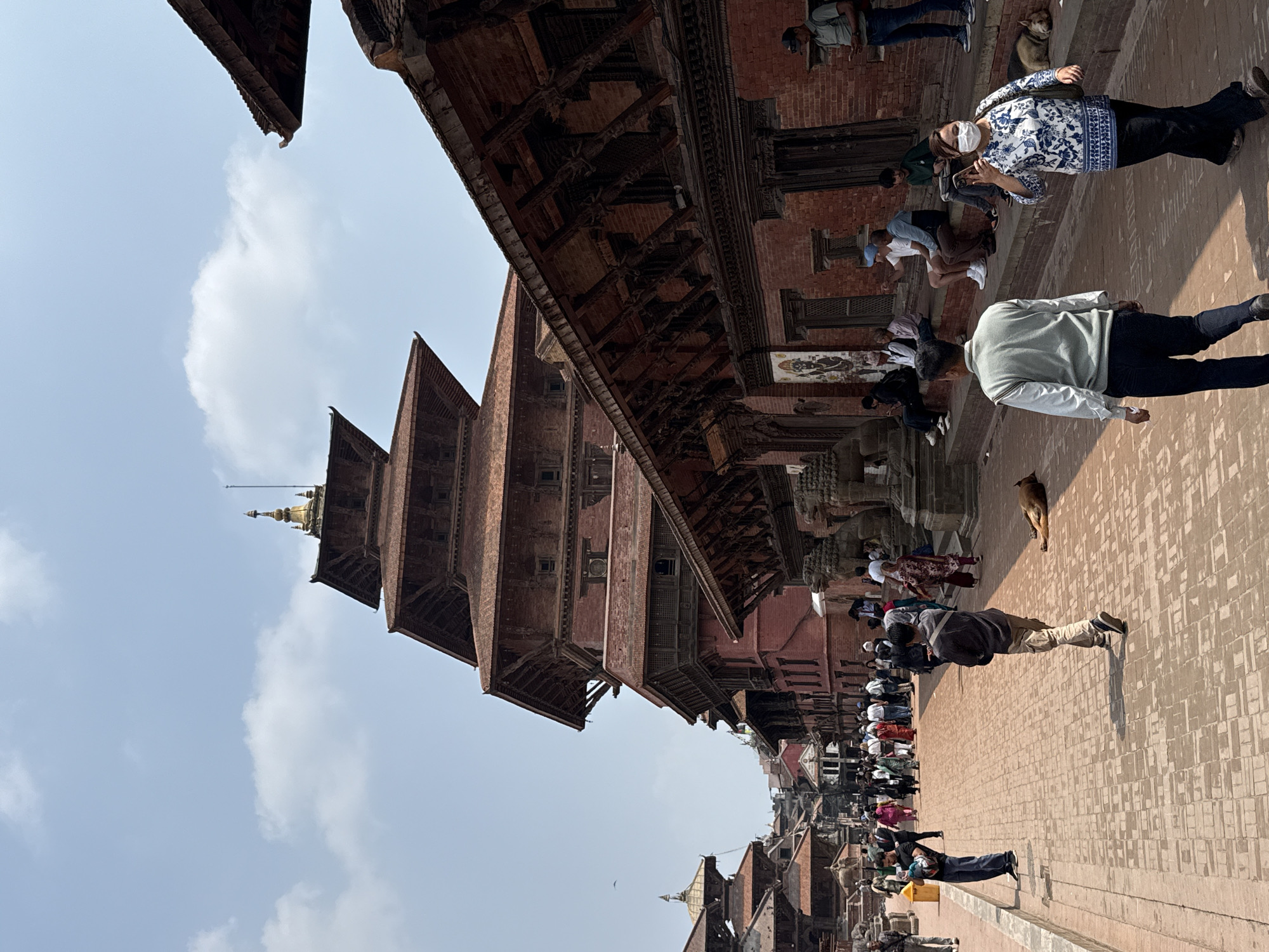 The image shows a tall, multi-tiered, reddish-brown wooden structure resembling a pagoda or temple, likely located in Durbar Square, Lalitpur, Nepal. The structure has a golden spire at the top and is set against a sky with scattered clouds. The foreground consists of a brick-paved plaza where people are walking. There are numerous individuals, including tourists. A dog also walks on the plaza. Many structures around the pagoda are made of brick with intricate wood carvings.
