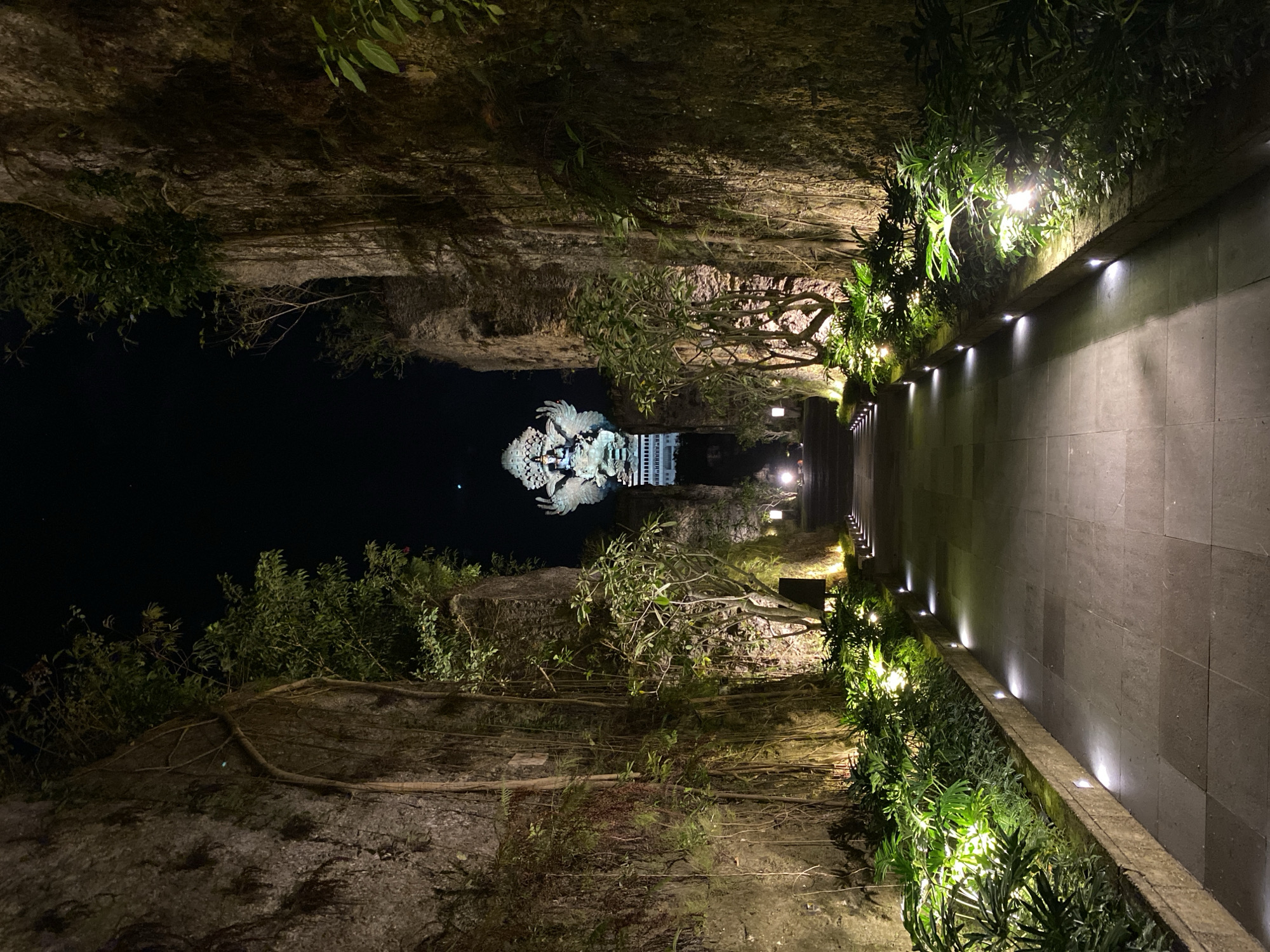 A long, paved pathway is illuminated by discrete lights embedded along its edges and in planter boxes lining the left side. The pathway leads towards a monumental statue of a deity with multiple arms, wings, and a headdress, which is lit from below. This statue stands in the distance at the end of the path, framed by towering, rough-hewn rock walls on either side. These walls are adorned with hanging vines and various plants, creating a natural, jungle-like atmosphere. The scene is set at night, with a dark sky above and soft, warm lighting highlighting the pathway, foliage, and the prominent statue. The location appears to be a sculpted natural environment, possibly an outdoor attraction or religious site.