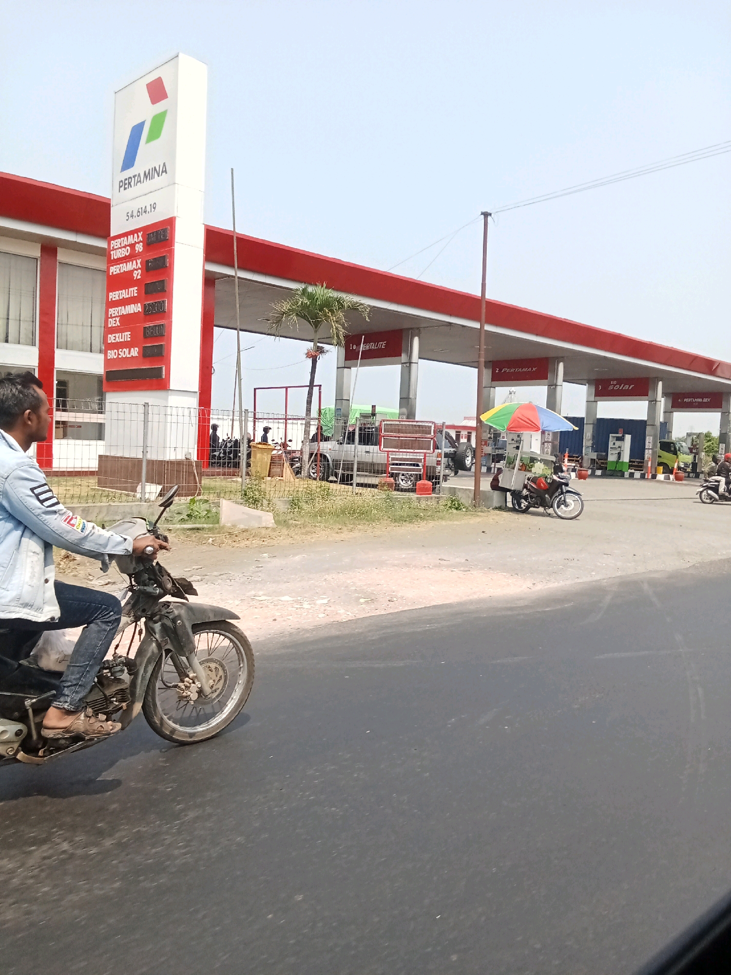The image depicts a street scene in Cangkringrandu, Indonesia, focused on a Pertamina gas station. In the foreground, a person on a motorcycle is riding on an asphalt road. The motorcycle is dark-colored and appears to be a standard model. The rider is wearing a light blue denim jacket and dark pants. Behind the motorcycle, the Pertamina gas station is visible with its distinctive red and white structure. A tall sign displays the Pertamina logo and fuel prices. Multiple fuel pumps are lined up under the covered station. In the middle ground, there are other vehicles, including a white truck and a parked motorcycle with a colorful umbrella over it, suggesting a small vendor. Several people are seen in the background near the gas station. The environment appears to be sunny, with a clear sky. A palm tree is also visible near the gas station. The overall scene captures a slice of everyday life at a roadside fuel stop in Indonesia.