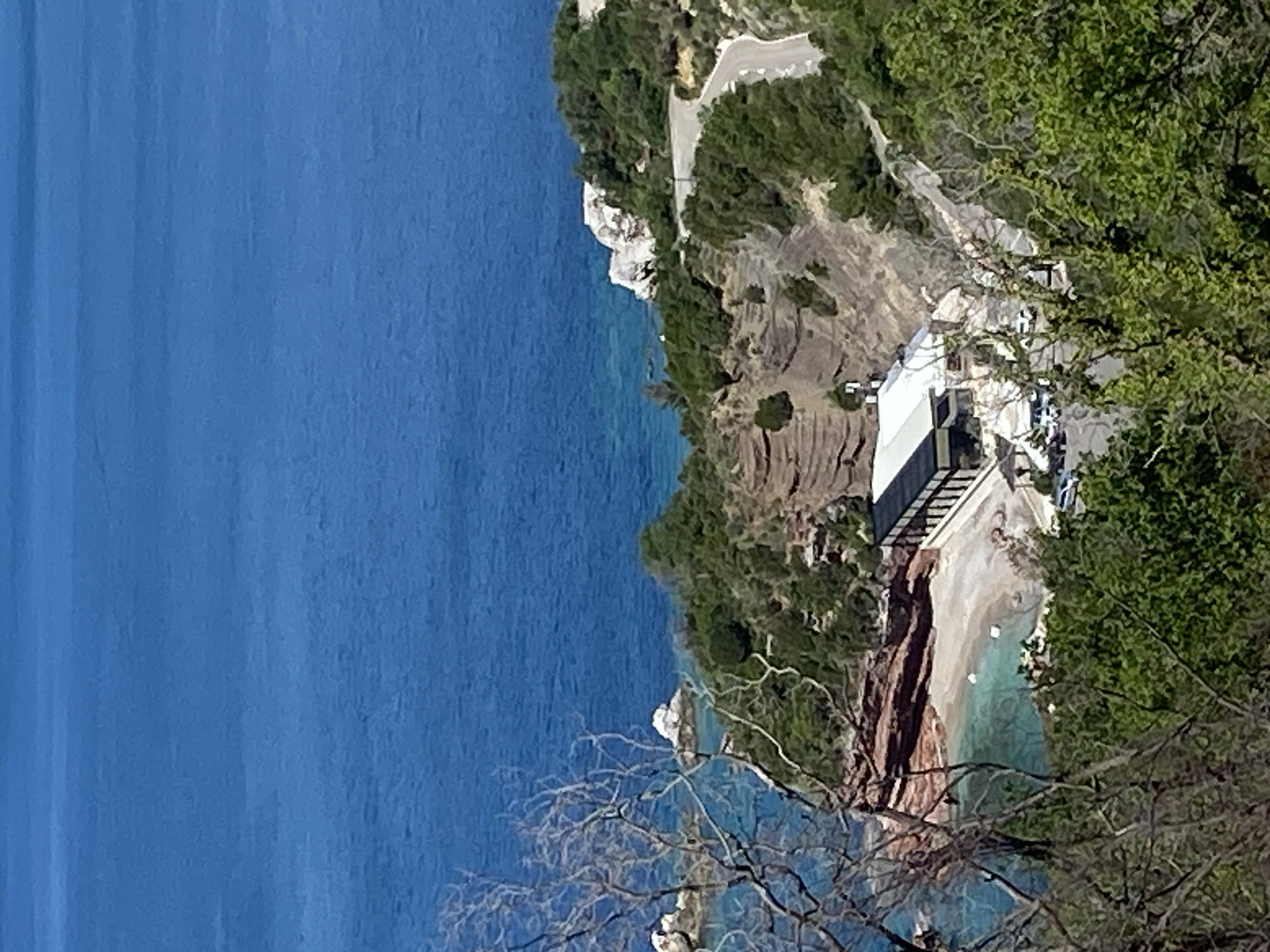 A tilted aerial view of a coastal scene in Blizikuće, Montenegro. A vibrant blue body of water dominates the left. A rugged coastline with cliffs covered in lush greenery and exposed rock formations runs along the right side. A winding road traces the edge of the cliff. Closer to the foreground, a modern building with a white roof is situated on a beach. A set of stairs leads down to the beach. The beach is composed of light-colored sand. The foreground includes a tree with bare branches.