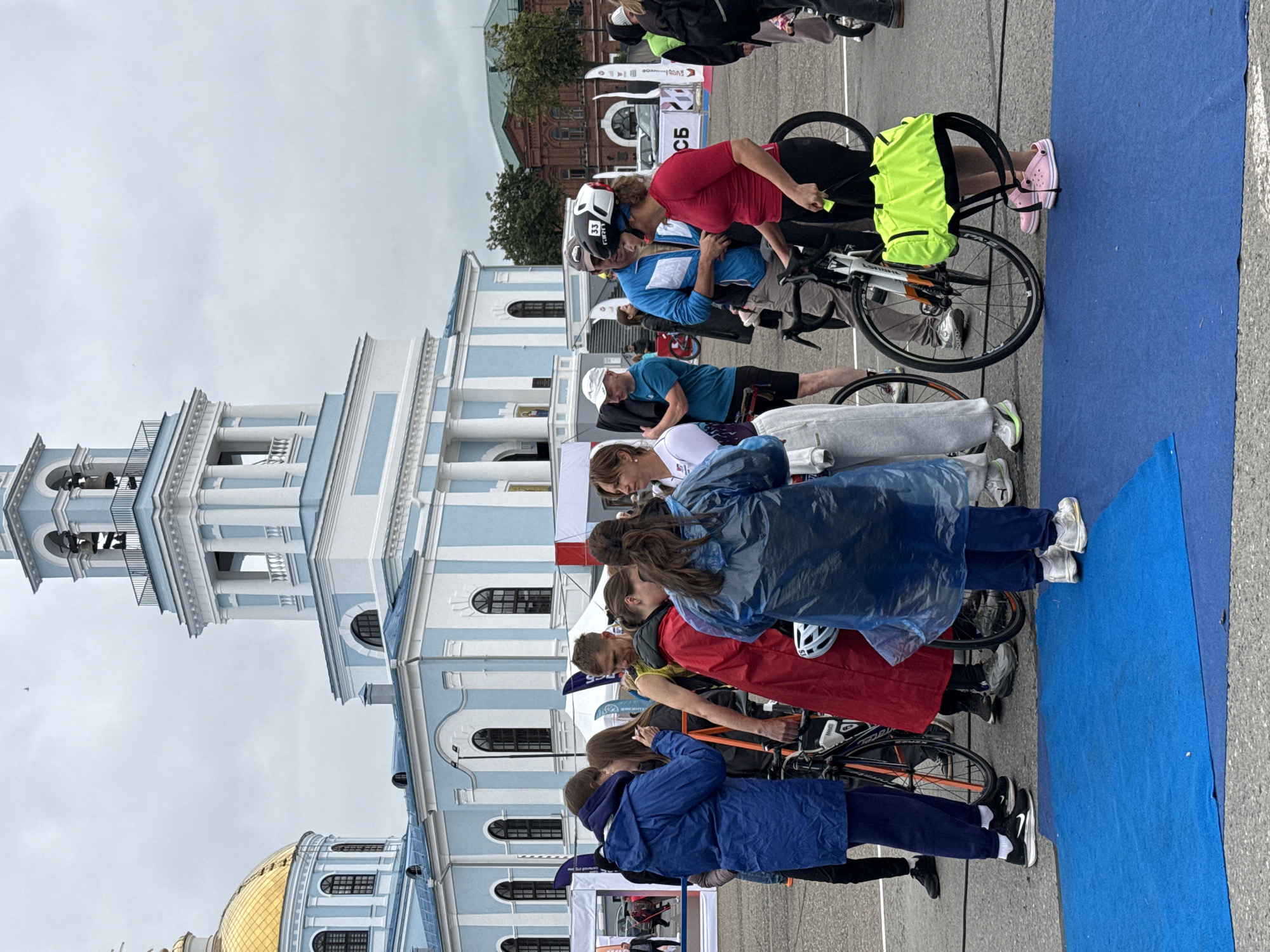 The image depicts a group of people gathered in an outdoor urban setting, likely before or after an athletic event such as a triathlon. In the background, a prominent blue and white building with classical architectural features and a golden dome is visible, identified as the Holy Trinity Cathedral in Votkinsk, Russia. Several individuals are standing, some with bicycles, suggesting participation in a cycling event. One woman in the foreground is wearing a red shirt and black pants, with a bicycle helmet on her head, and holding a bright yellow sports bag. Another person in a blue raincoat is standing next to a bicycle. The ground is paved, and a blue mat is laid out. The sky is overcast, indicating cloudy weather. Flags and banners are also present, suggesting an organized event. The overall atmosphere appears to be one of casual gathering and preparation or debriefing.