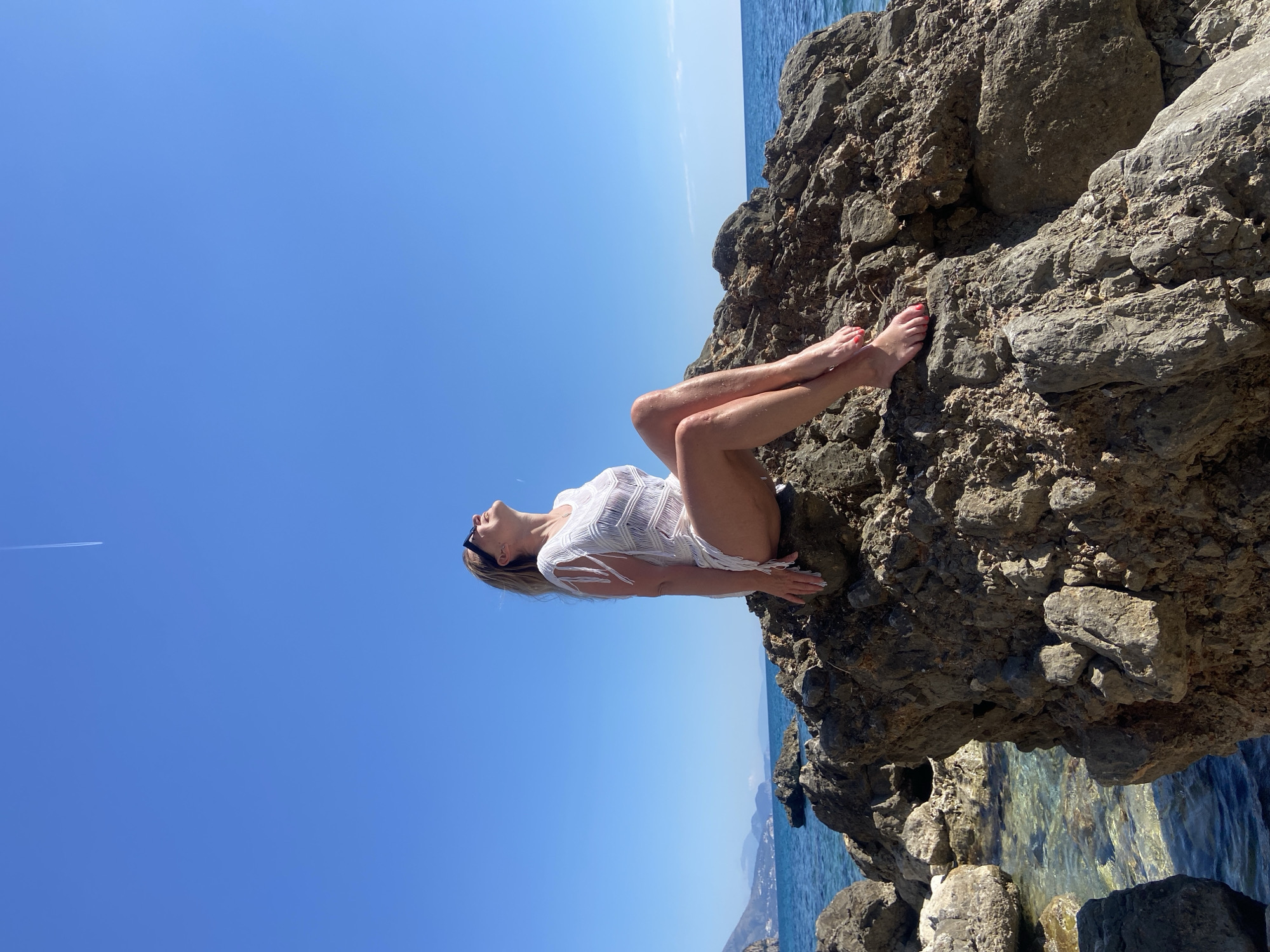 A woman wearing a white cover-up and sunglasses is reclining on a rocky outcrop in Prijevor, Montenegro. The rocks are weathered and dark, with clear turquoise water visible beneath them. The sky is a bright, cloudless blue. There is a plane contrail visible in the upper left corner of the image. The terrain is rugged coastline. The woman is facing upwards with her legs crossed at the ankles, resting on the rocks.