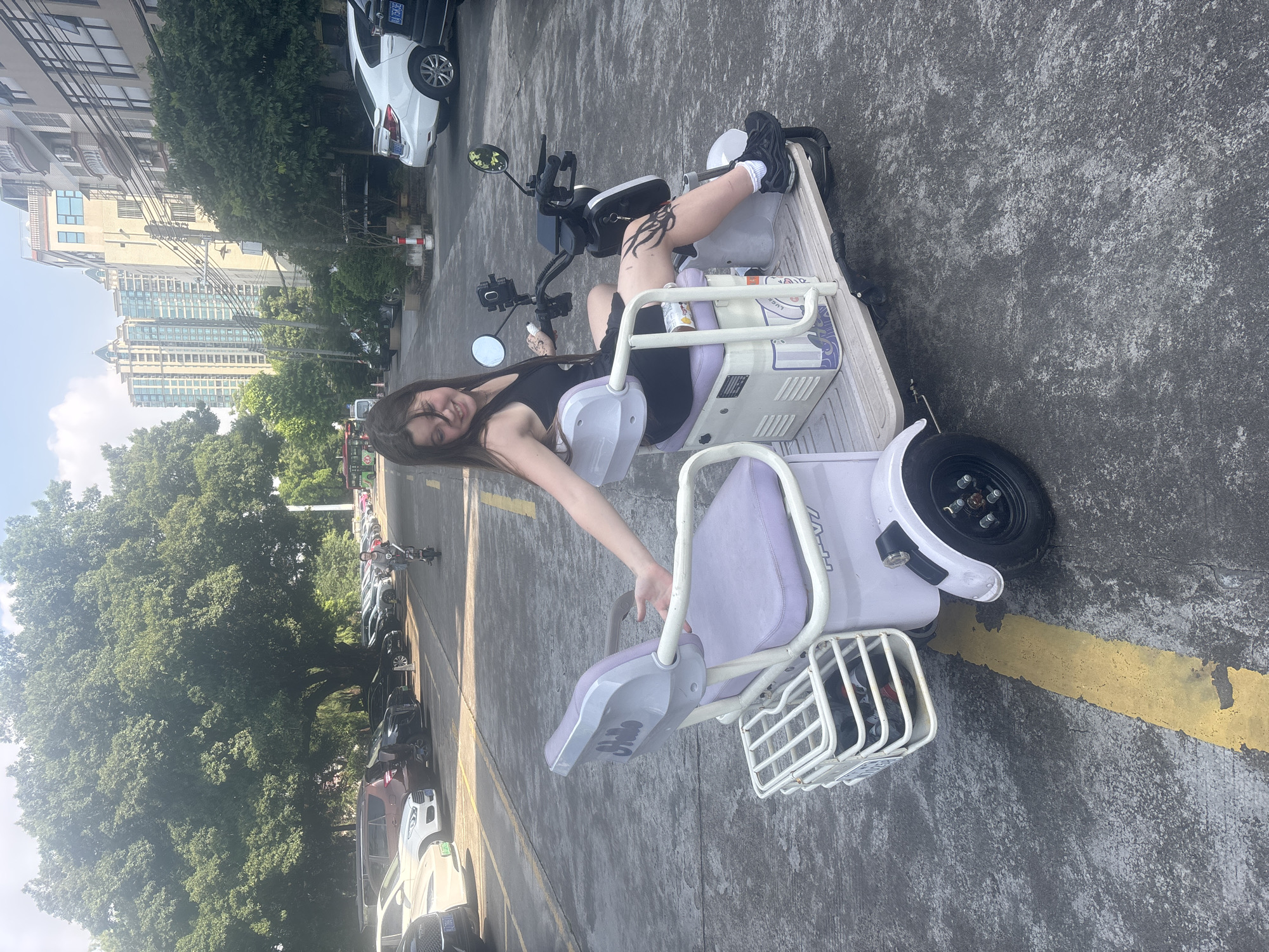 A young woman with long brown hair, wearing a black tank top and black athletic shoes, sits on a three-wheeled electric scooter with a purple seat in a parking lot. She is leaning back and smiling at the camera, with her left arm extended. She has a tattoo on her left thigh. The scooter is white and purple, with a white basket on the front and a white storage compartment behind the seat. In the background, there are parked cars, trees, and buildings. The scene appears to be in Nanhai District, China, given the presence of buildings and vehicles common to urban China. A yellow line is painted on the asphalt ground.