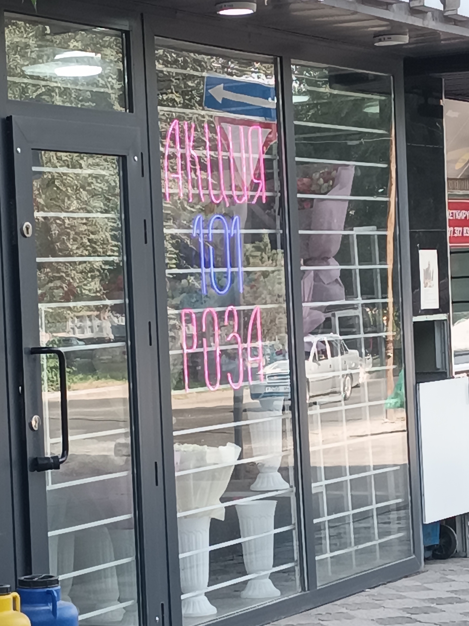 The image shows the storefront of a flower shop with a large glass window. Neon signs are visible inside the store, displaying the words "АКЦИЯ 101 РОЗА" (Promotion 101 Roses). Reflections in the glass reveal trees and a street with a white car driving by. Inside, white vases and what appear to be flower arrangements are displayed. The entrance to the store is on the left side of the image, with a dark metal door and frame. The flooring outside the shop is a checkered pattern of light and dark grey tiles. This appears to be a commercial establishment in a city environment.