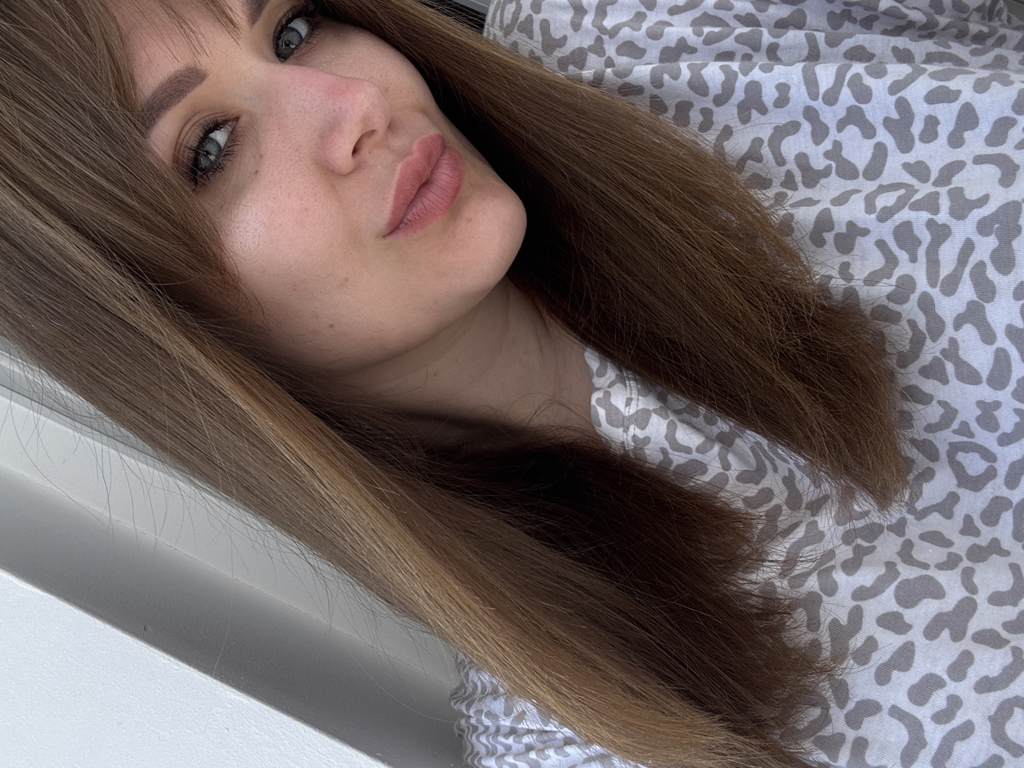 A close-up photograph shows a woman with fair skin and brown hair. She is wearing a gray and white patterned shirt. The location is not discernible from the image. The photograph primarily focuses on the woman's face and hair.