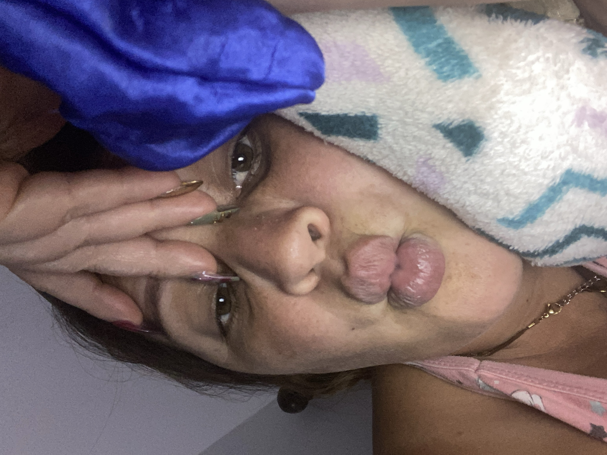 A close-up, low-angle indoor shot shows a woman with dark hair and brown eyes puckering her lips. Her right hand is positioned on her forehead, with long, dark nails visible. A blue satin cloth is draped behind her head, and a white towel with purple and teal geometric patterns is placed on her cheek. She is wearing a pink tank top with a floral print and a gold necklace. The lighting is dim, suggesting the photo was taken in a bedroom or similar private space, likely at night or with the lights off. There are no discernible urban or city landmarks in the image.