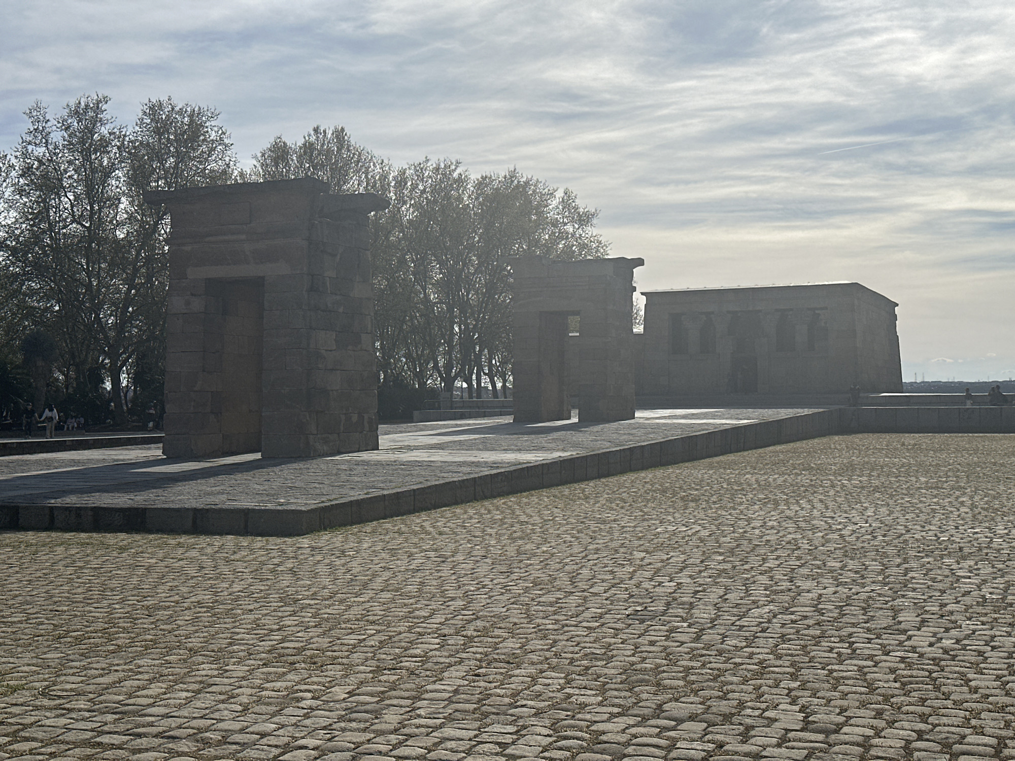 The image shows the Temple of Debod, located in Madrid, Spain. The temple is situated on a raised platform paved with stone. The foreground consists of a cobblestone plaza. Behind the temple, trees are visible against a cloudy sky. Several people are present in the background.