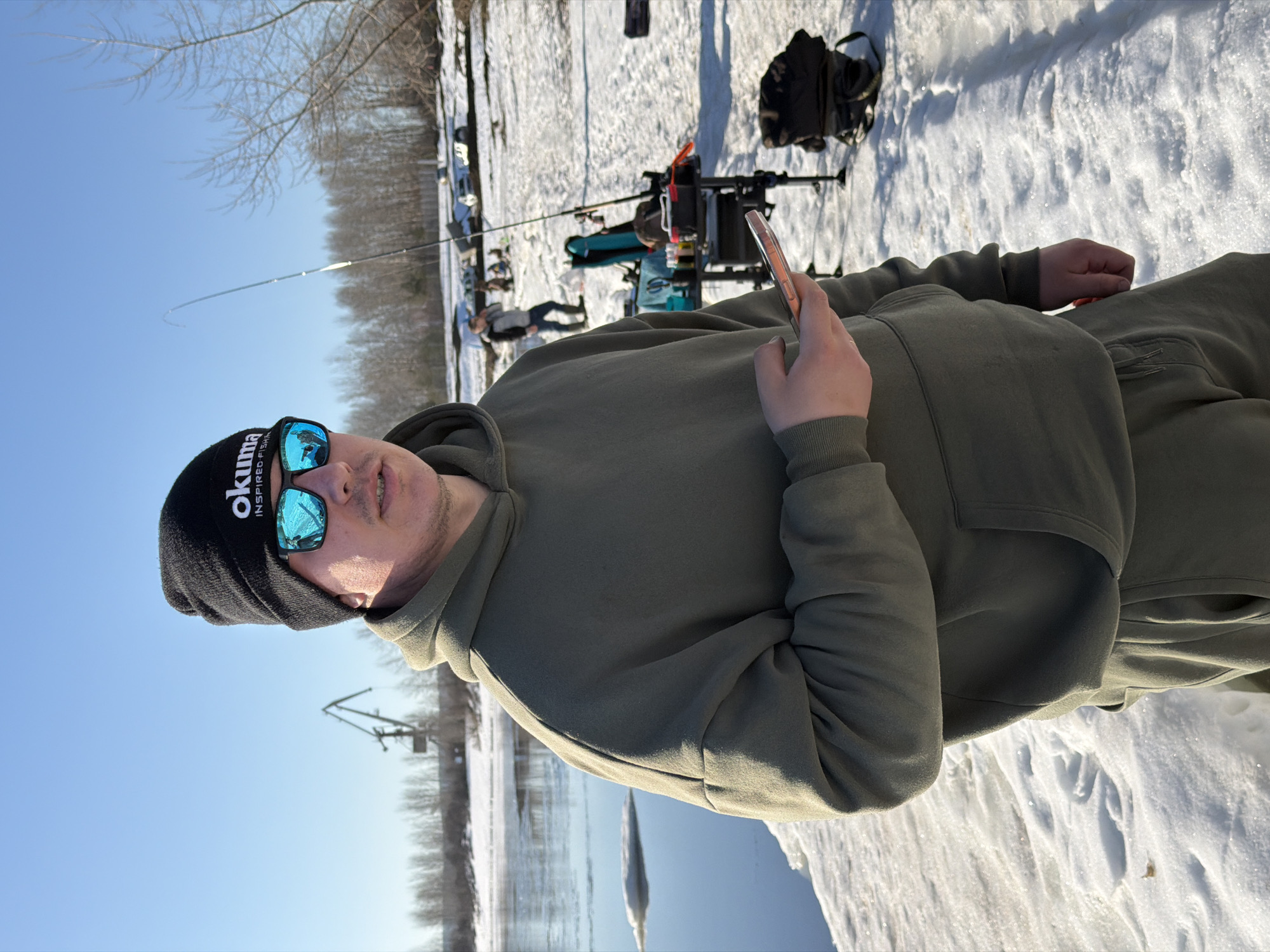 A medium, horizontally-oriented, outdoor shot shows a man standing in a snowy field along a body of water. The man is dressed in a drab olive green hooded sweatshirt and pants. He is wearing a black knit beanie with "Okuma" printed on it and reflective blue sunglasses. He holds a silver smartphone in his left hand. In the background, several fishing poles are set up near a tackle box. The environment includes snow-covered ground, bare trees, and a blue sky. It appears that it may be close to a small body of water as well. According to information online, this picture was taken in the city of Chaikovsky, Russia.
