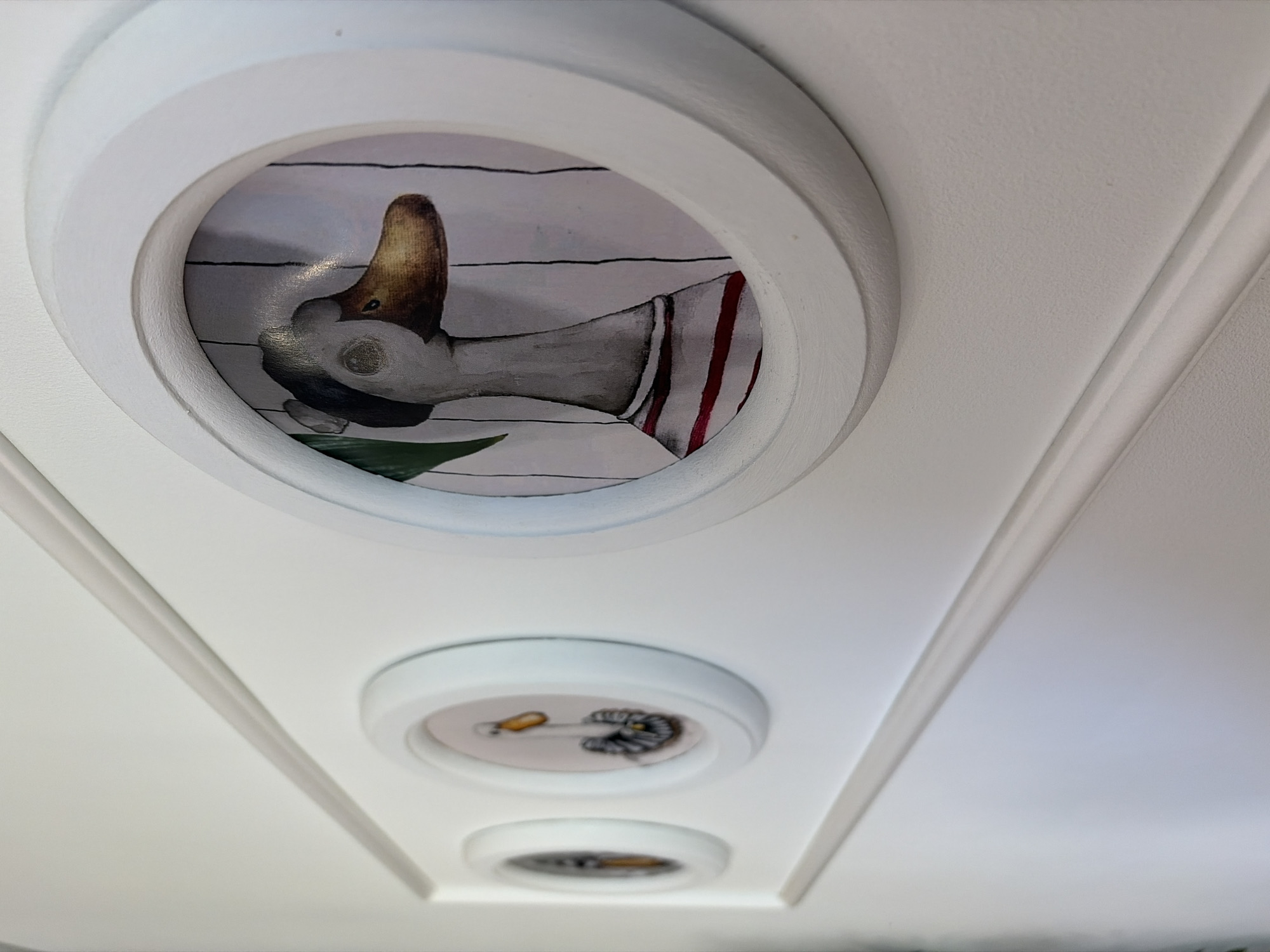 A close-up, angled shot shows a section of a white ceiling with decorative molding. Three circular insets are visible, each containing a painting. The largest, at the top, features a close-up of a duck's head and neck. The duck has a brown and yellow bill, grey and white plumage, and appears to be wearing a striped red and white garment. The background of this painting is a series of horizontal white lines.
Below this, two smaller circular insets are visible. The middle inset contains a painting of what appears to be a drill bit with a yellow handle, and some metallic gears or components. The bottom inset is out of focus, but seems to contain a similar image with a yellow element. The ceiling itself is white and textured, with white molding that creates a framed effect around the insets and in long, linear channels. The lighting suggests it is daytime, and the overall impression is one of domestic decoration.