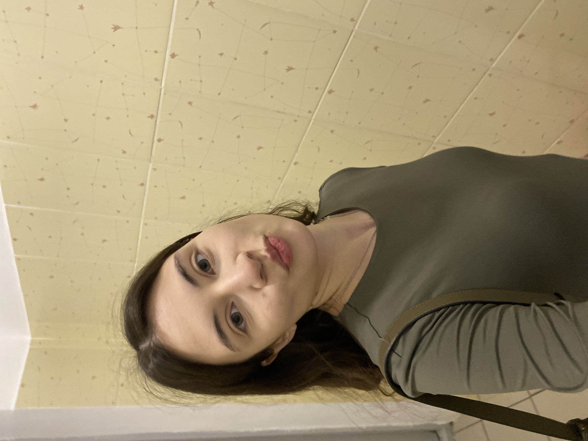 A young woman with fair skin, dark brown hair, and green eyes is looking up at the camera. She is wearing a long-sleeved, olive green shirt, and a strap (likely from a purse) is visible across her torso. The background consists of a wall covered in light yellow tiles with a constellation-like pattern in brown. The bottom left of the image shows a portion of a white corner or edge, possibly a doorway or window frame. The location is likely a restroom or kitchen, given the tilework. There is no information in the image that would specifically identify the city as Votkinsk, Russia. The description provided assumes the location provided is accurate but not visible in the image.
