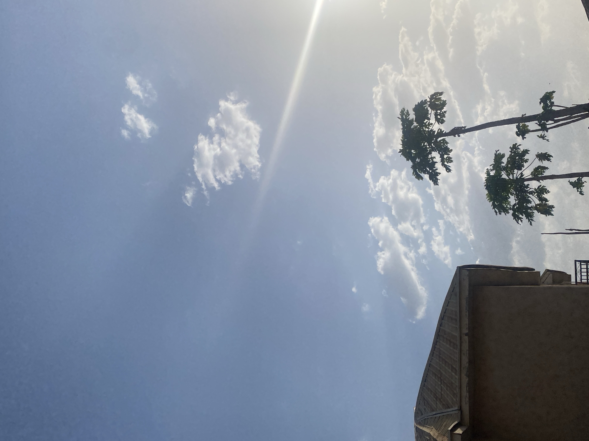 The image displays an outdoor scene in Sabon Tasha, Nigeria. The sky occupies the majority of the frame, exhibiting a clear blue hue with scattered white cumulus clouds. The sun is visible in the upper right quadrant, casting a prominent diagonal light ray across the sky. In the upper left portion of the image, the top section of a building is visible, featuring a multi-faceted, corrugated metal roof that appears weathered and rusty-brown. A portion of the building's light brown exterior wall is also present. On the right side of the frame, several tall, slender trees, identifiable as papaya trees by their characteristic trunks and clusters of leaves at the apex, stand against the sky. No individuals or specific actions are depicted. The scene is illuminated by daylight.
