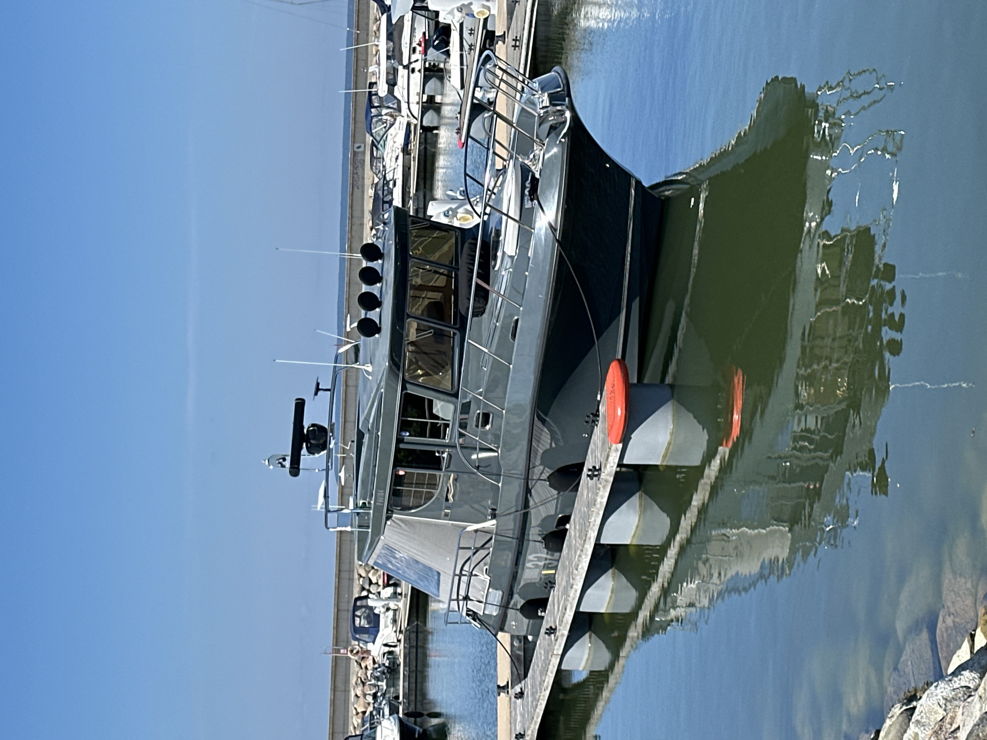 The image shows a dark grey motor yacht docked at a pier in a marina on a clear day. The yacht has a covered cabin with windows and a radar unit on top. There are other boats docked nearby in the background. The water is calm and reflects the yacht and the sky. The marina appears to be located in Tallinn, Estonia, with a stone wall and some vegetation visible along the shoreline in the background. There are no people visible in the image. The scene is a typical view of a marina on a sunny day.